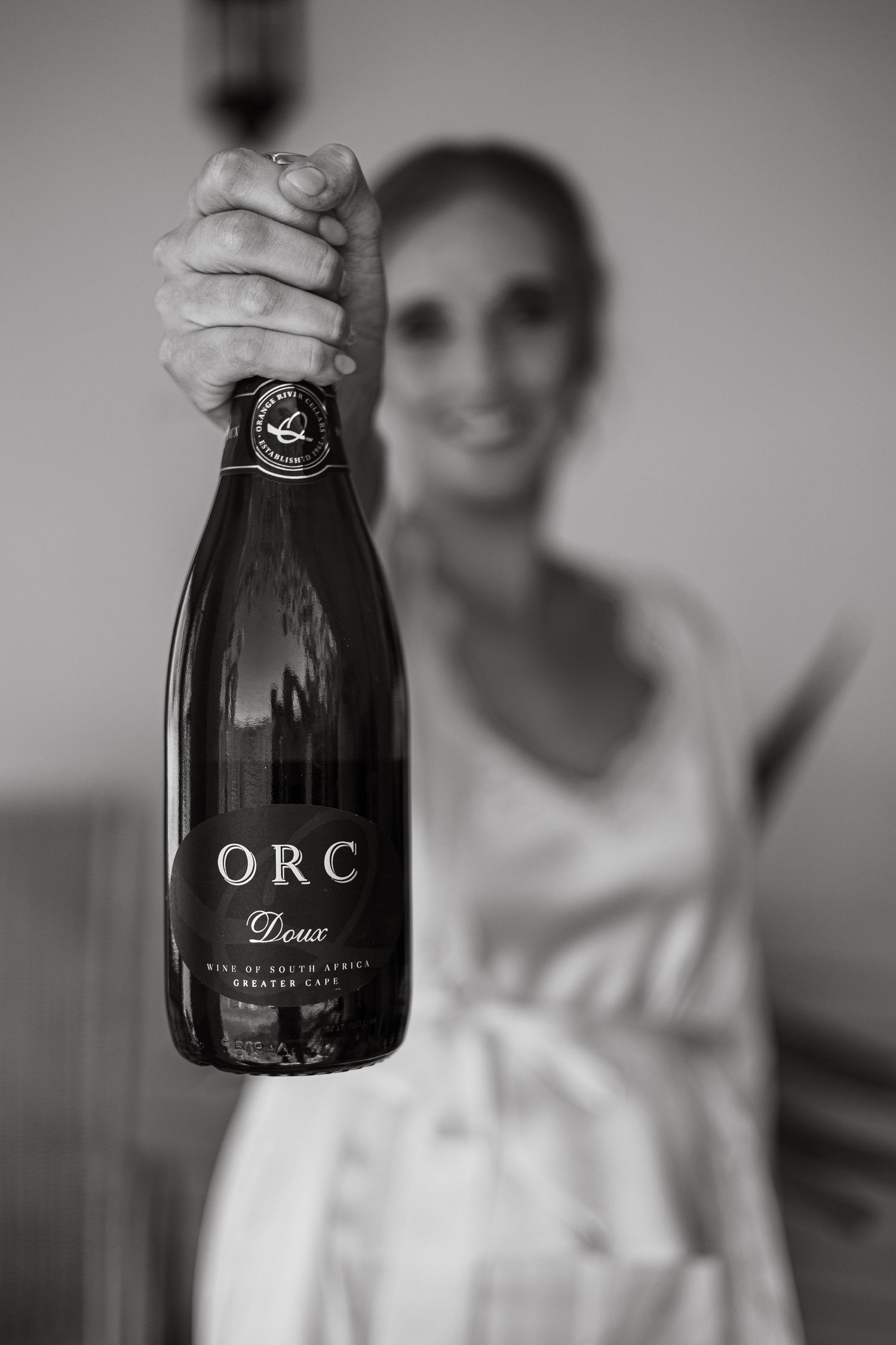 Black and white close-up of a smiling bride holding up a bottle of ORC Doux sparkling wine from South Africa.