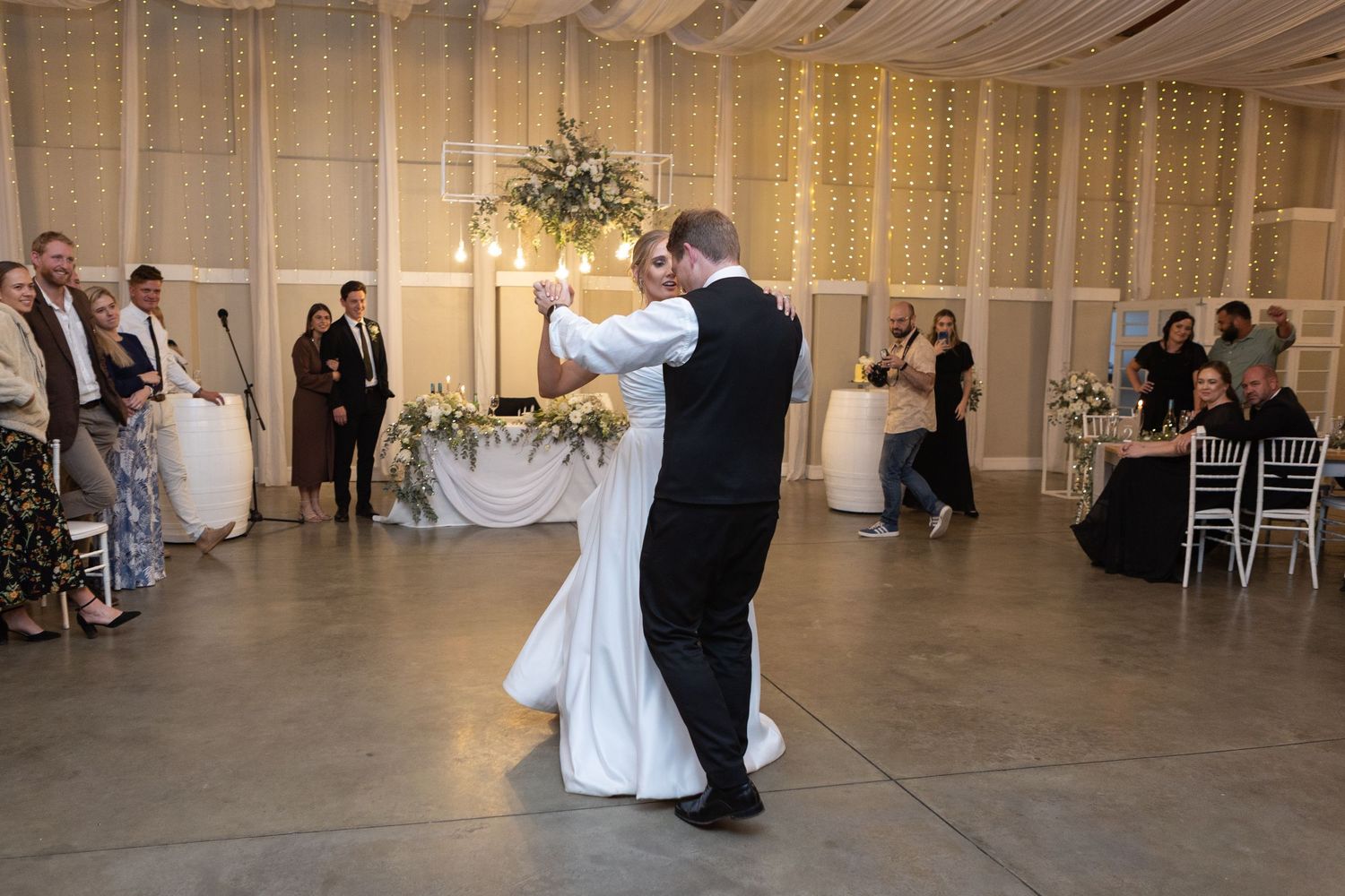 Bride and groom share their first dance at elegant wedding reception with string lights and floral arrangements.