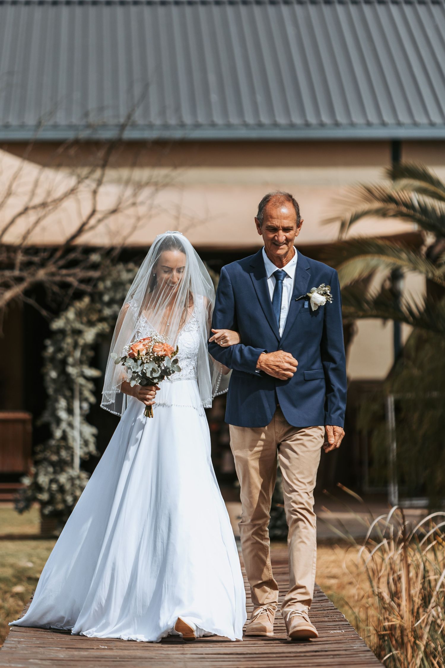A bride in a flowing white dress walks down a wooden path with a companion in a navy suit and khaki pants.