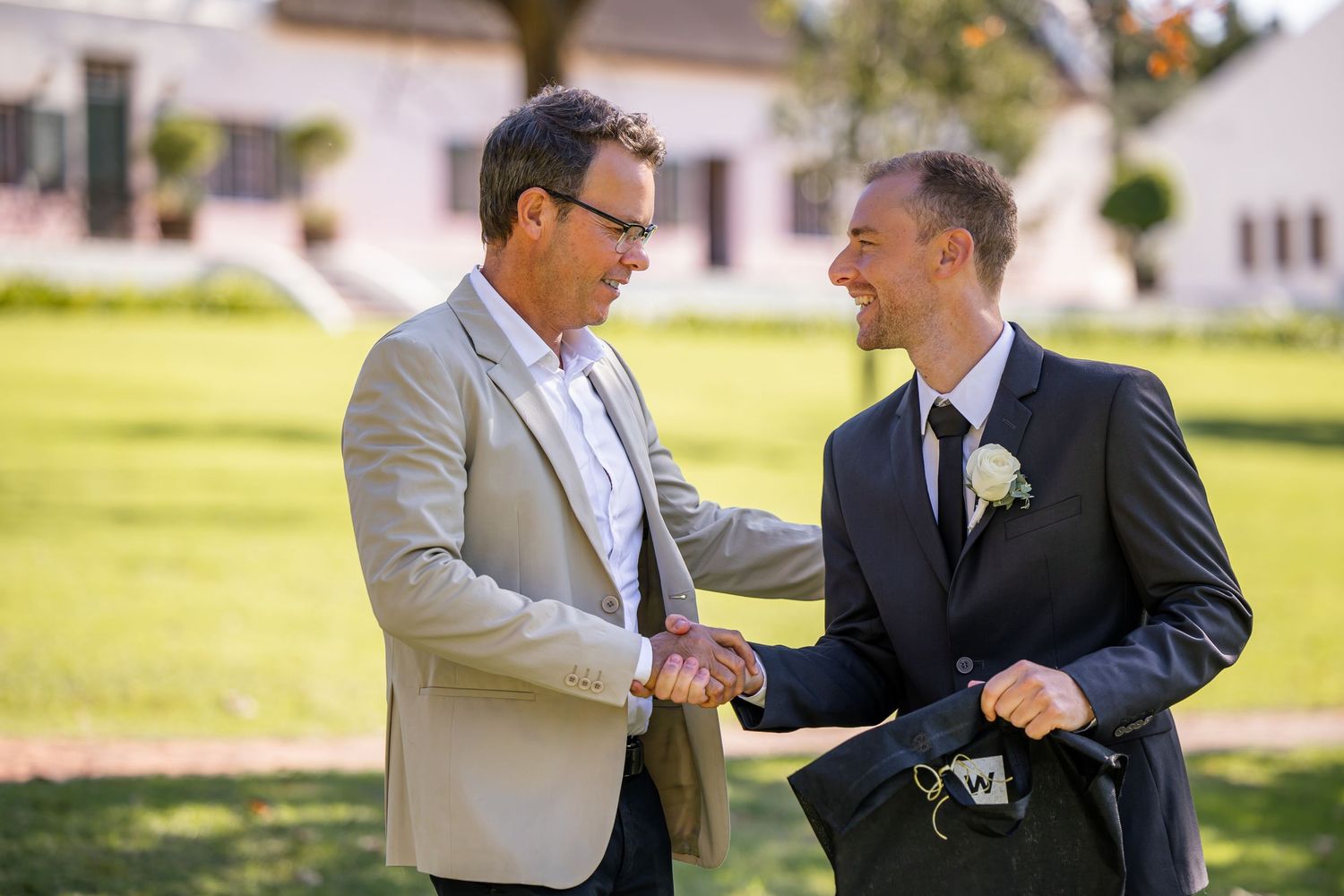 Two people in formal attire share a joyful fist bump greeting outdoors on a sunny day with houses in the background.