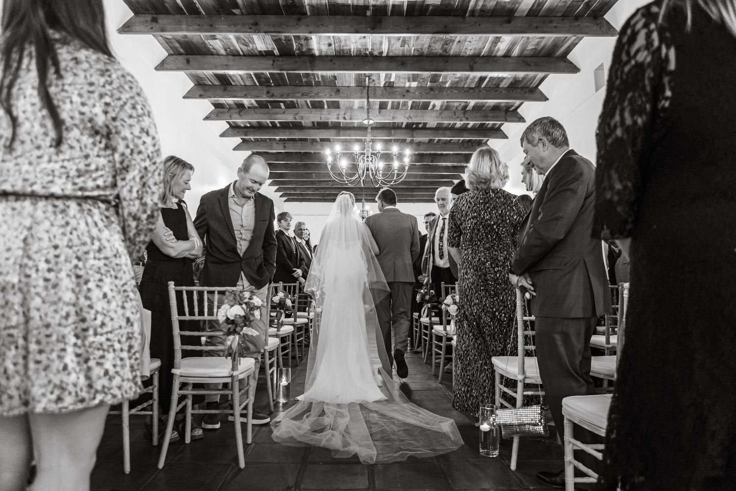 Black and white wedding ceremony photo showing bride walking down the aisle with guests standing in elegant indoor venue.