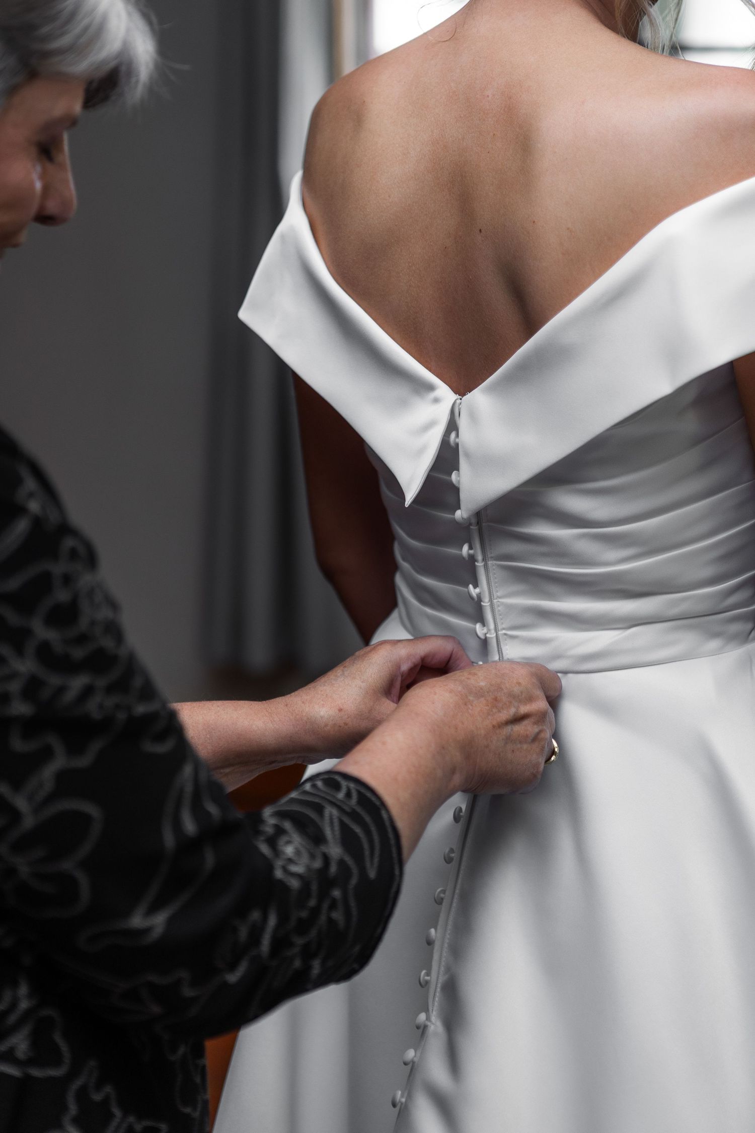 Mother carefully fastens buttons on the back of bride's off-shoulder white wedding gown.