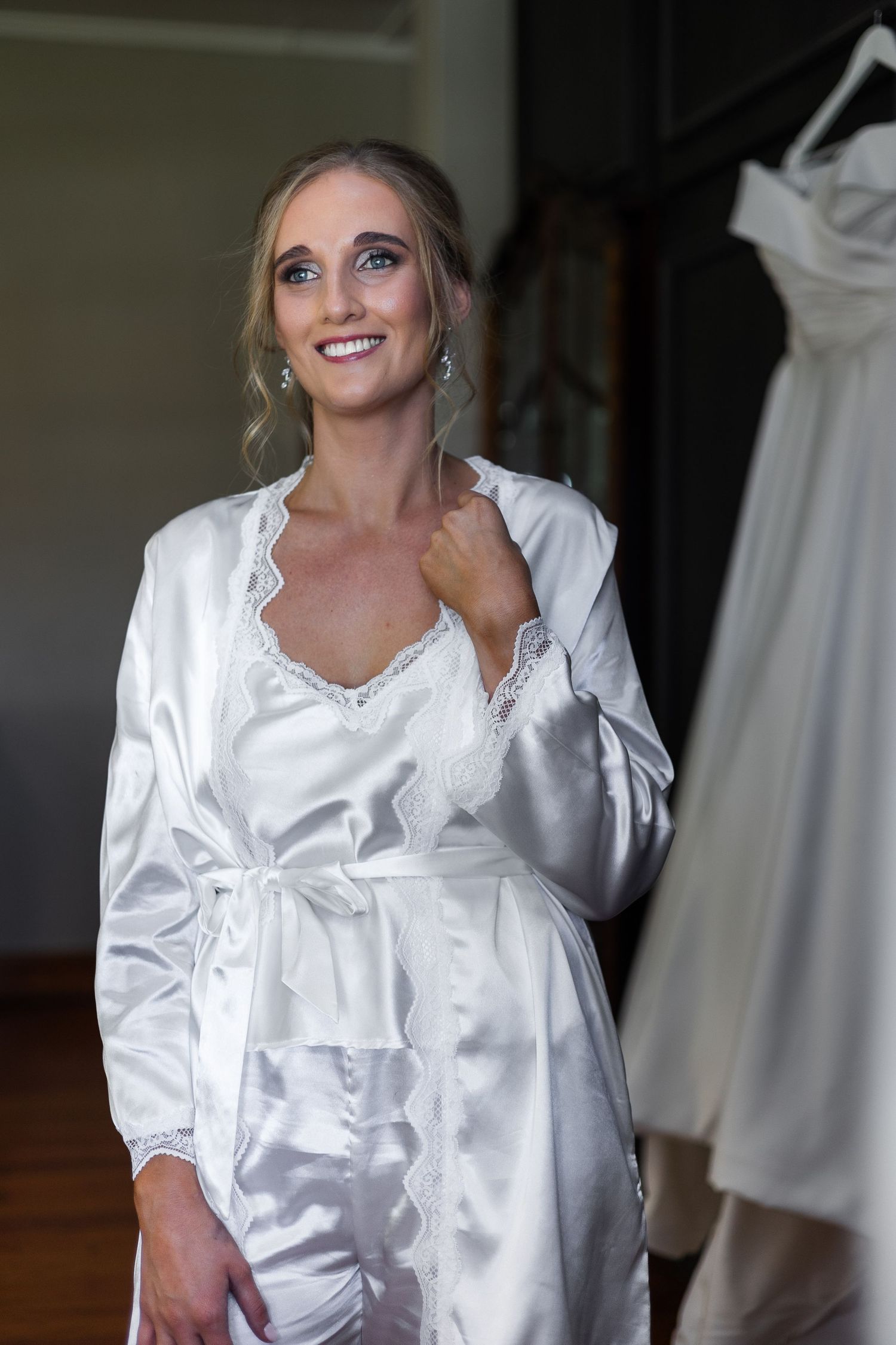 Smiling bride in white satin lace robe standing near her wedding dress before ceremony.