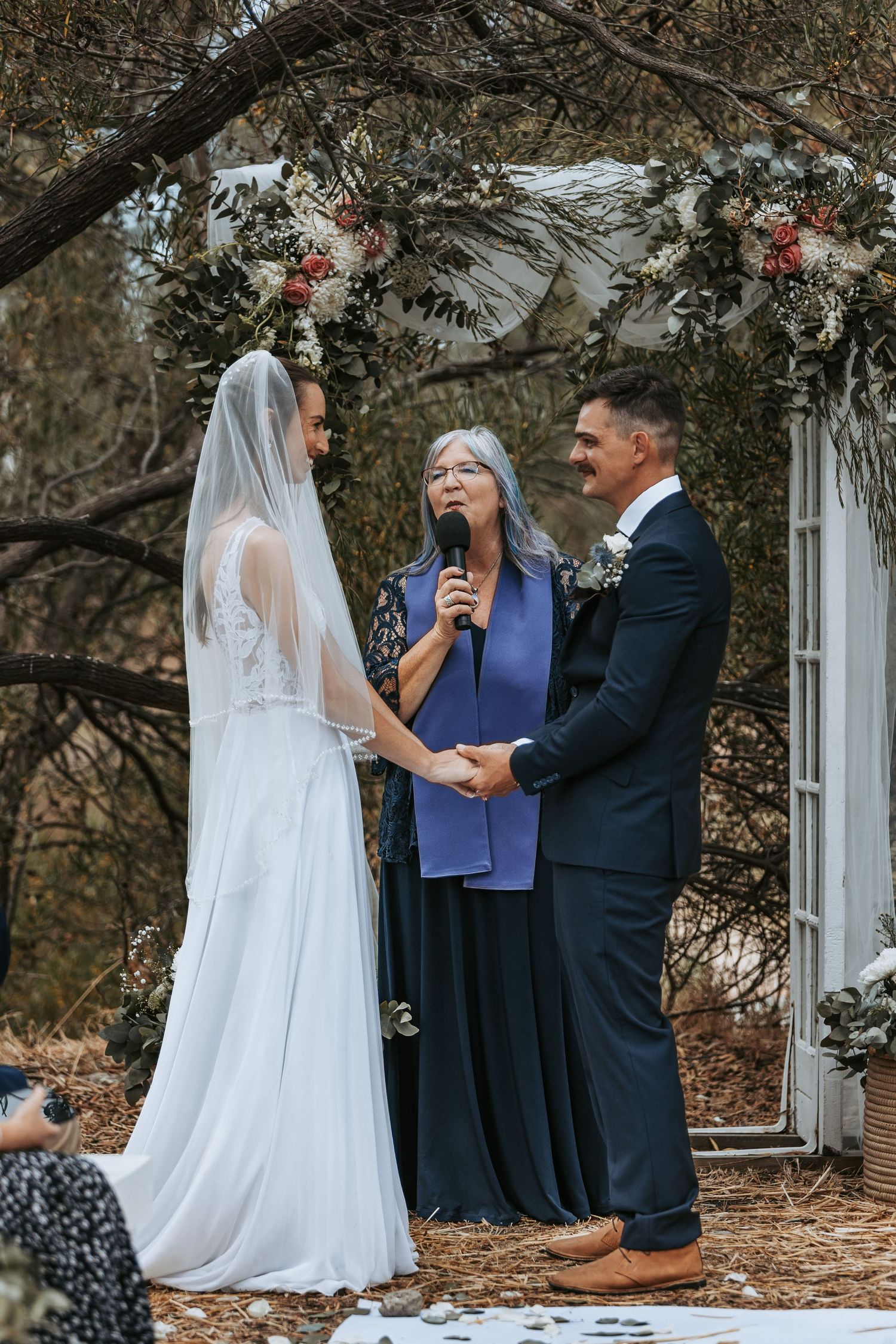 Wedding ceremony in progress under trees with officiant in blue and couple in formal attire.
