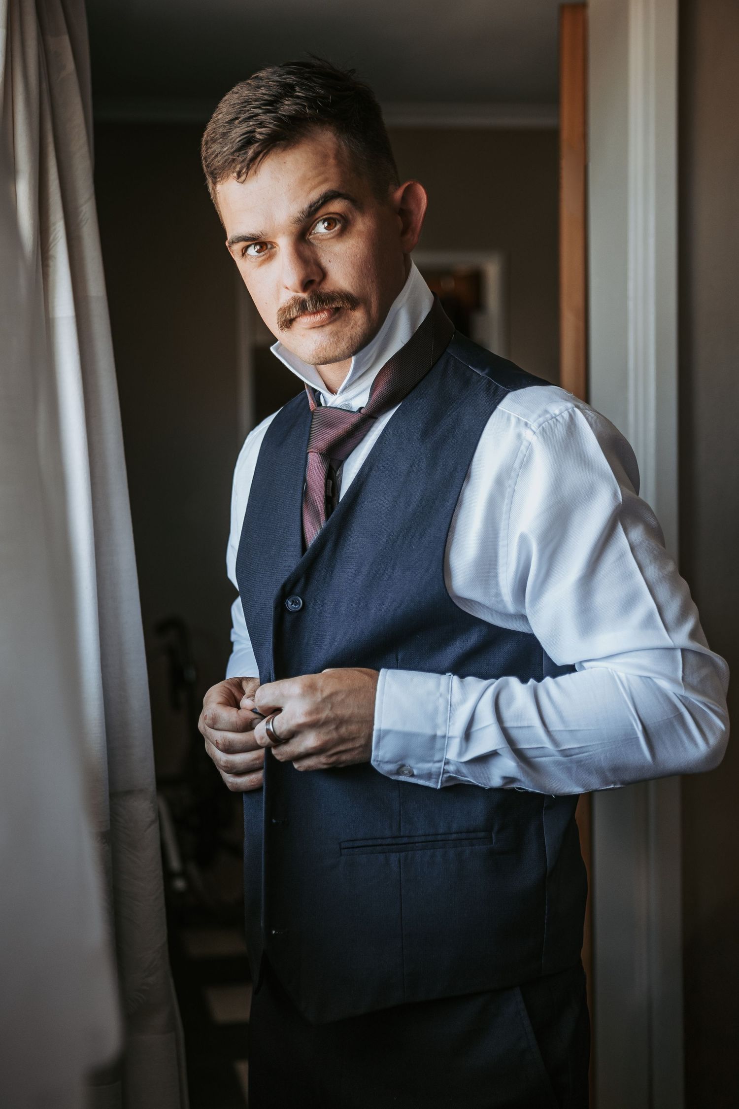 Stylish groom adjusts his vest while getting ready in a dimly lit room with natural window light.