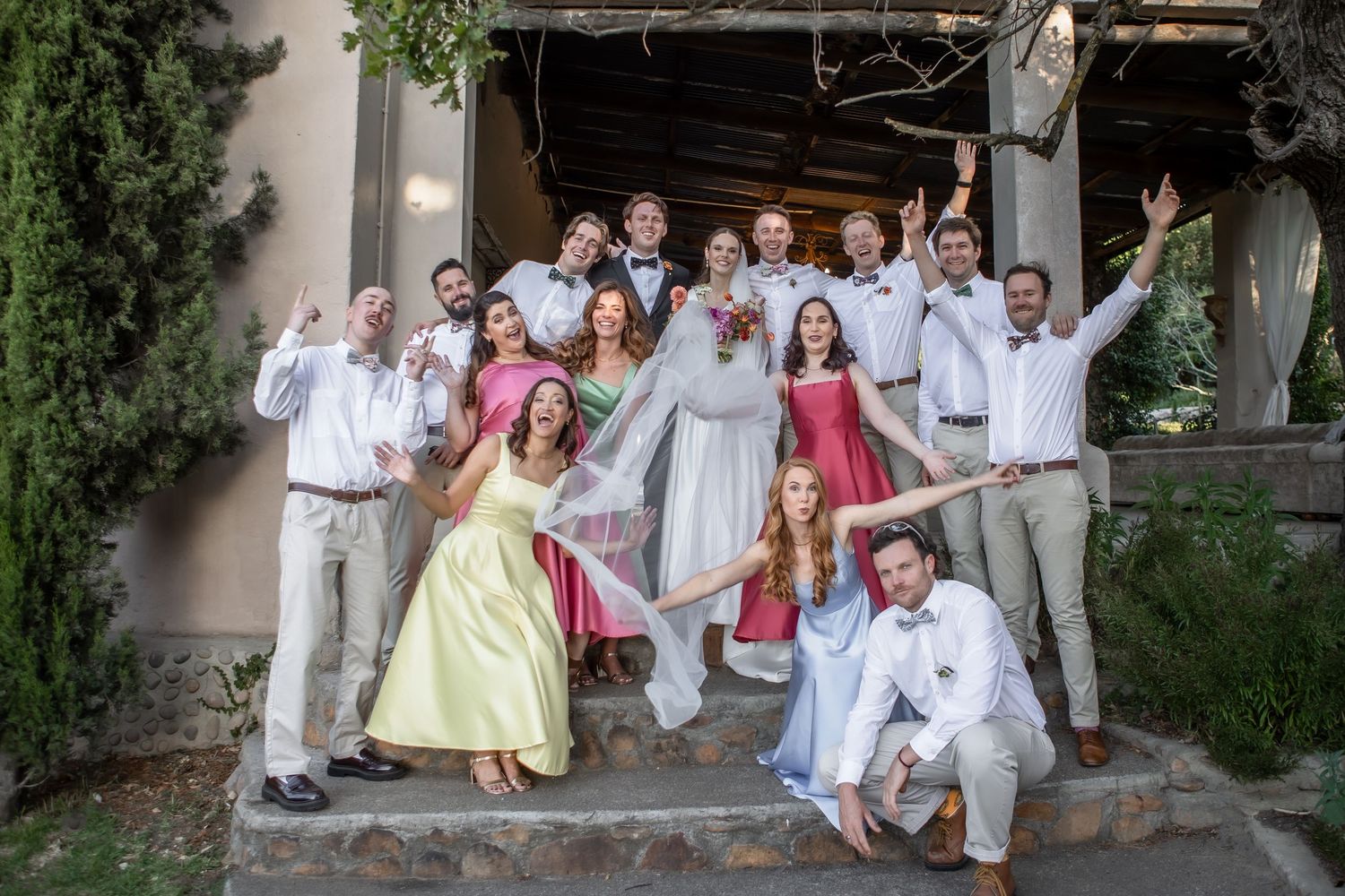 A wedding party poses on rustic steps with bridesmaids in pastel dresses and groomsmen in light colored suits.