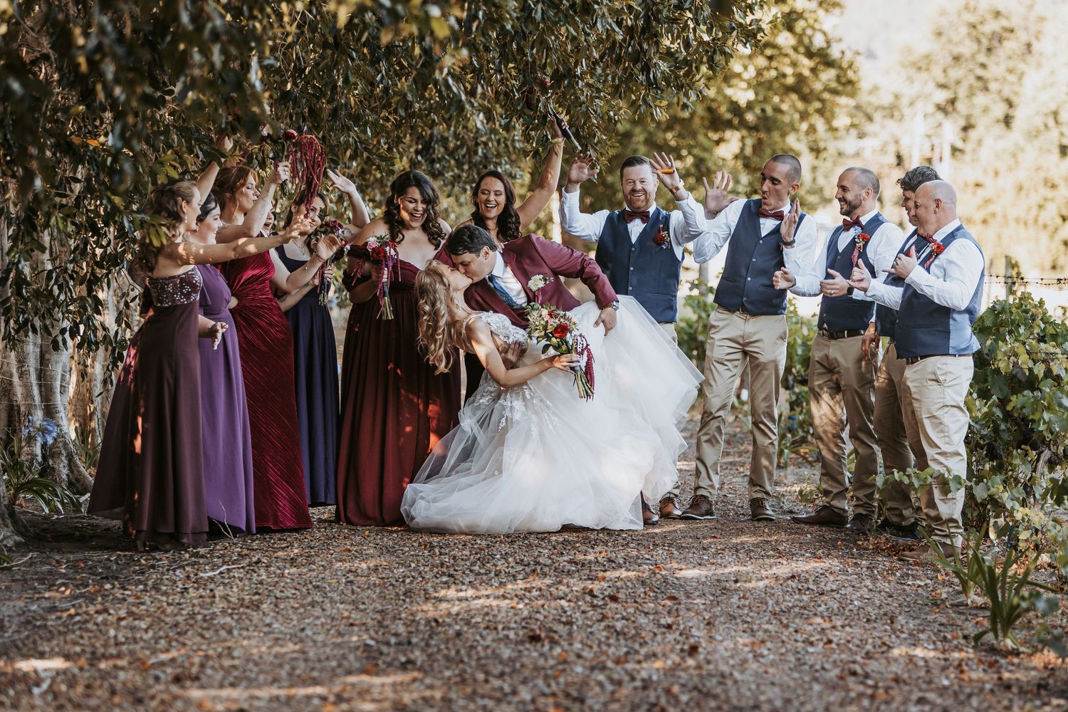 Wedding party members pose for photos outdoors under trees with natural lighting.