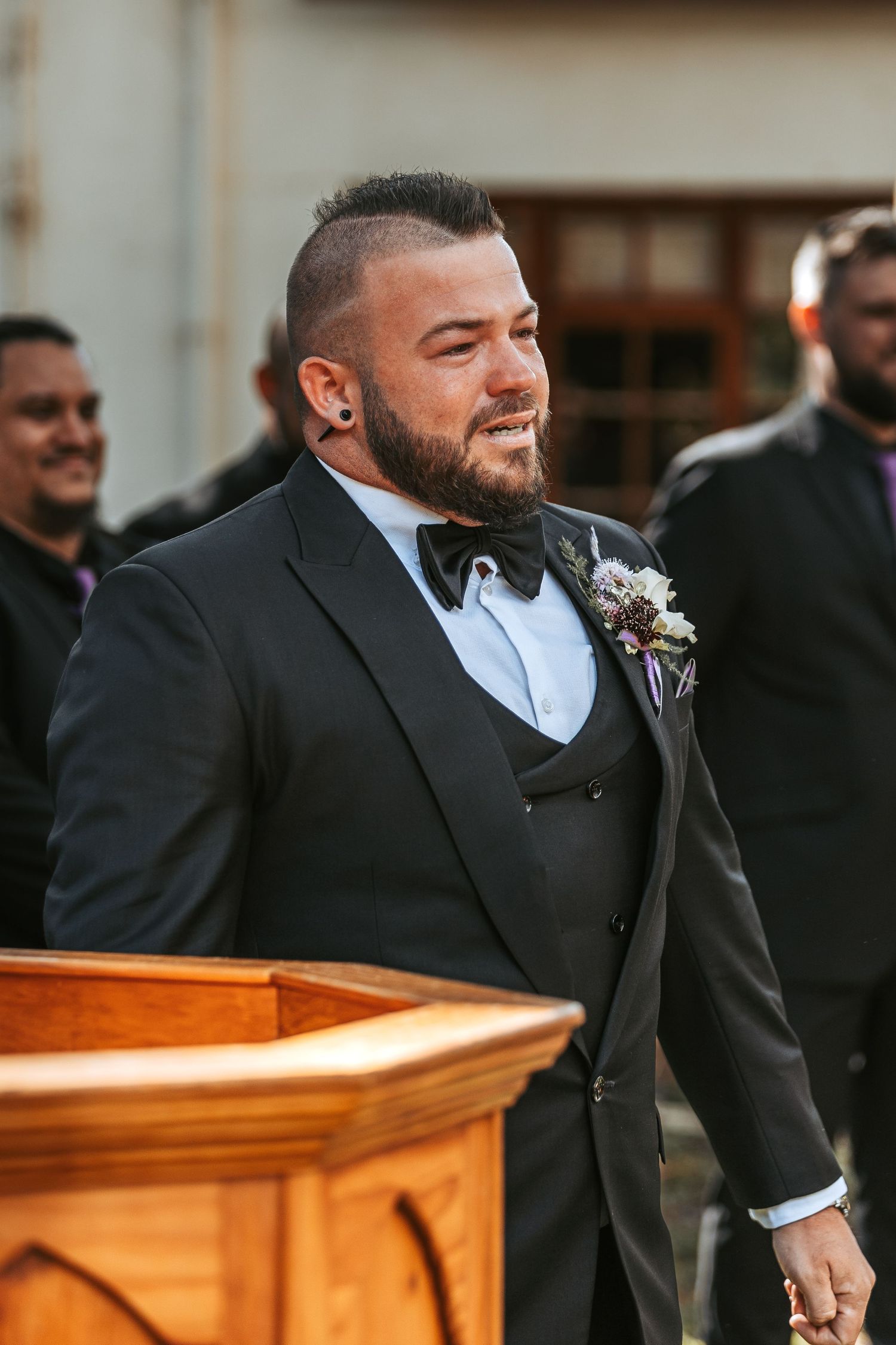 Groom in classic black tuxedo smiling during outdoor wedding ceremony.