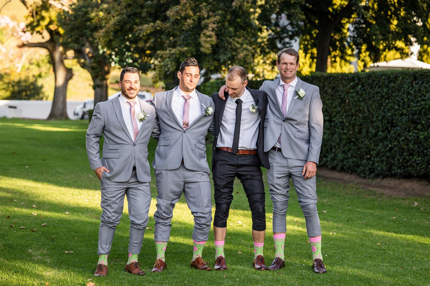 Groomsmen in gray suits pose together on a sunny lawn for wedding photos.