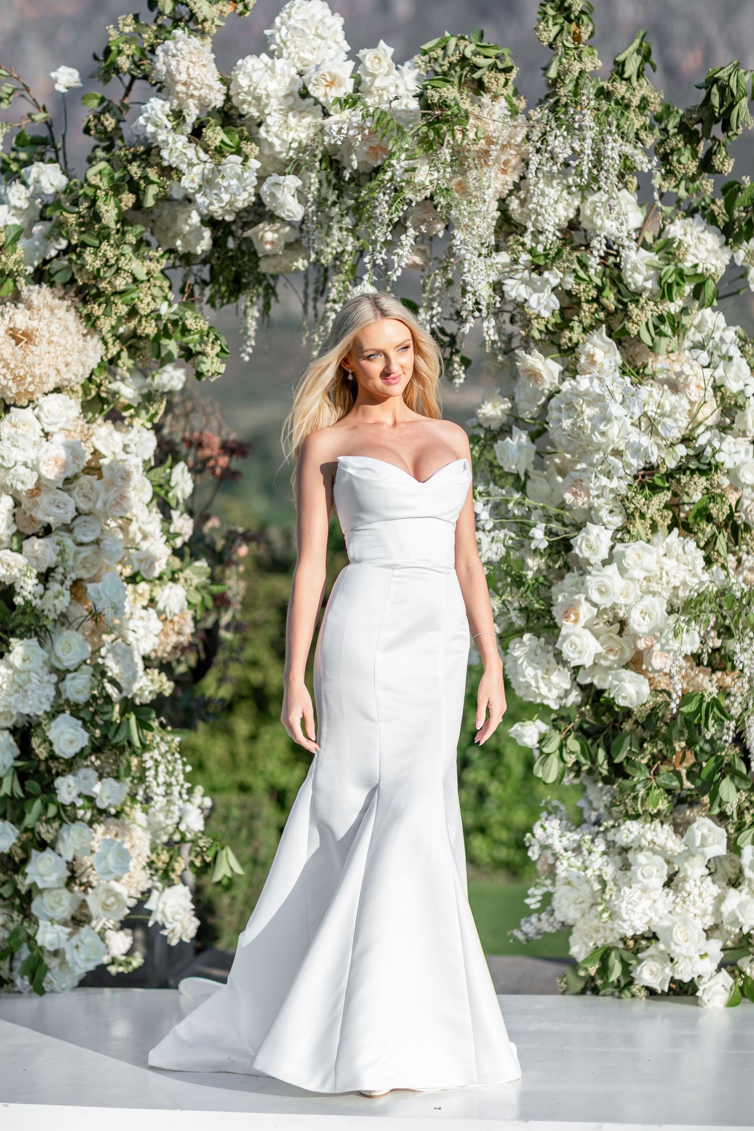 A bride in a strapless white wedding gown poses in front of a romantic floral arch with white roses and greenery.