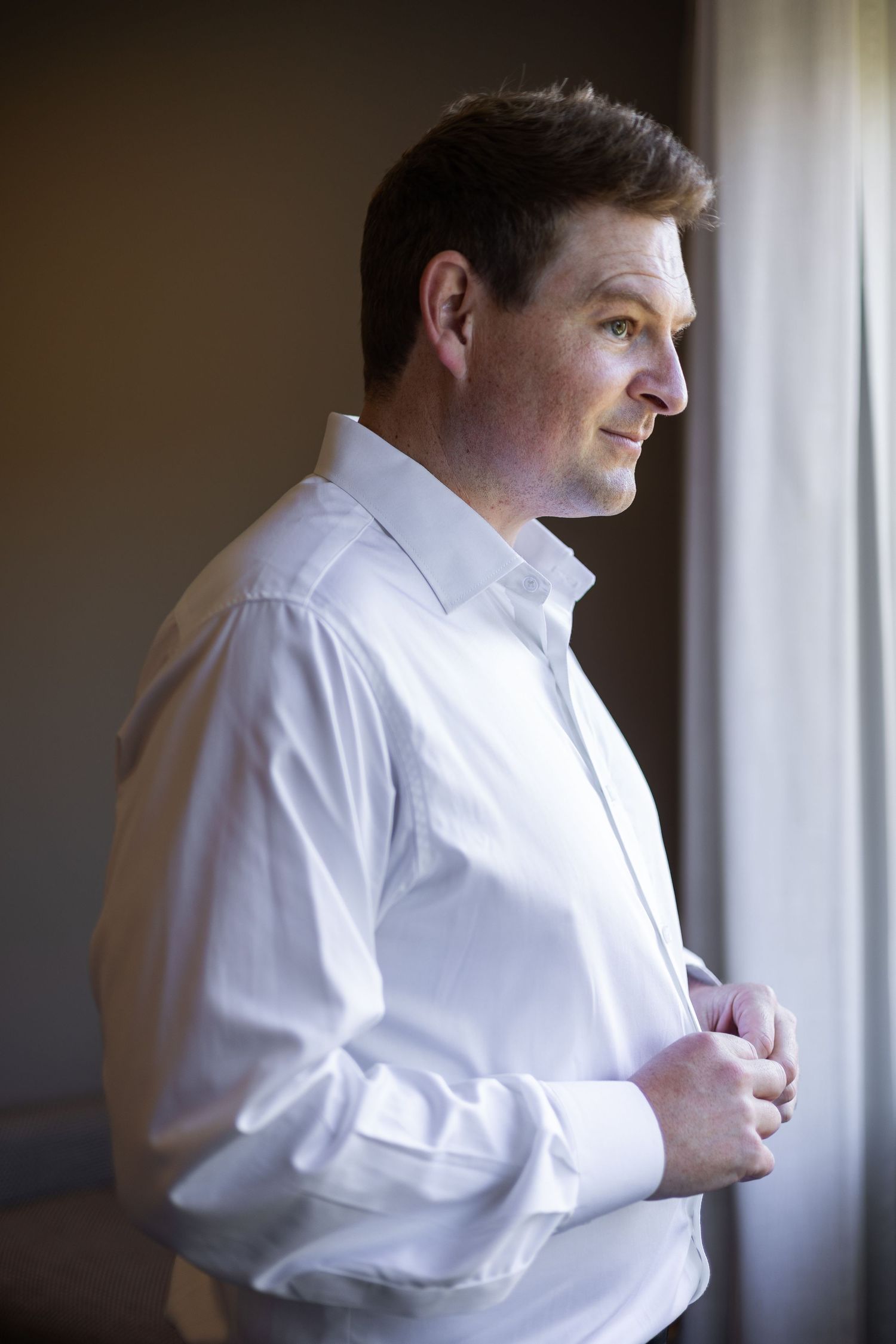 A man in a white dress shirt buttoning up near a window with soft natural light.