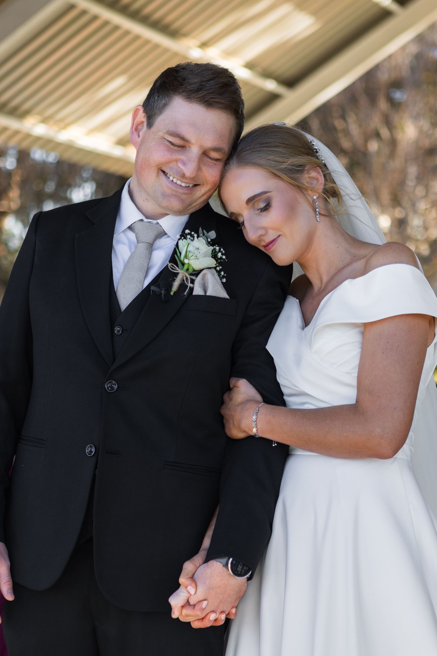 Newlywed couple holding hands and leaning together tenderly under a shaded outdoor structure.