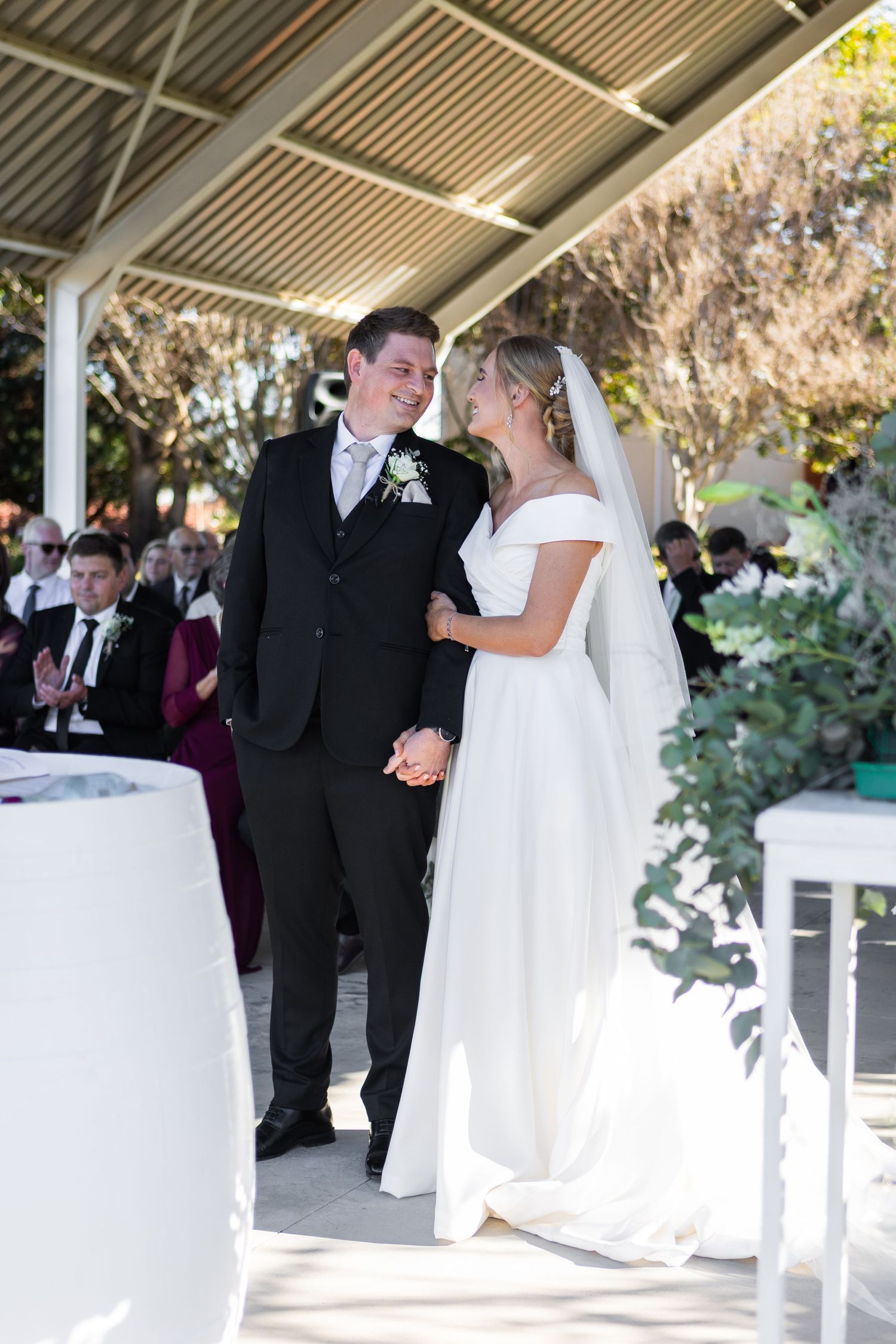 Bride and groom holding hands and smiling at outdoor wedding ceremony under pavilion with guests seated behind.