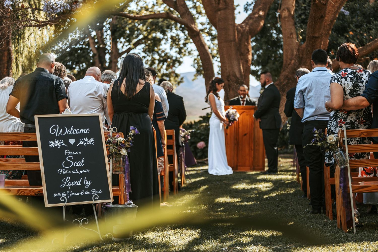 Wedding ceremony setup outdoors with chalkboard signs and wooden chairs under trees.