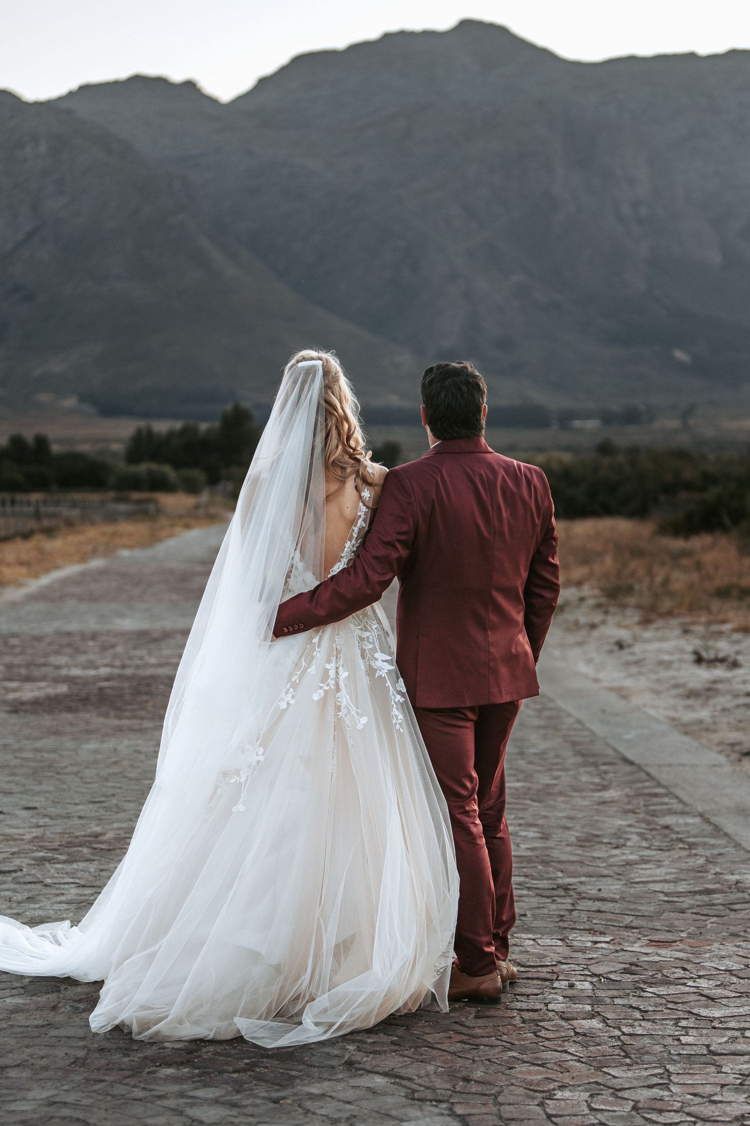 A couple in wedding attire stands against a dramatic mountain backdrop on a cracked desert ground.