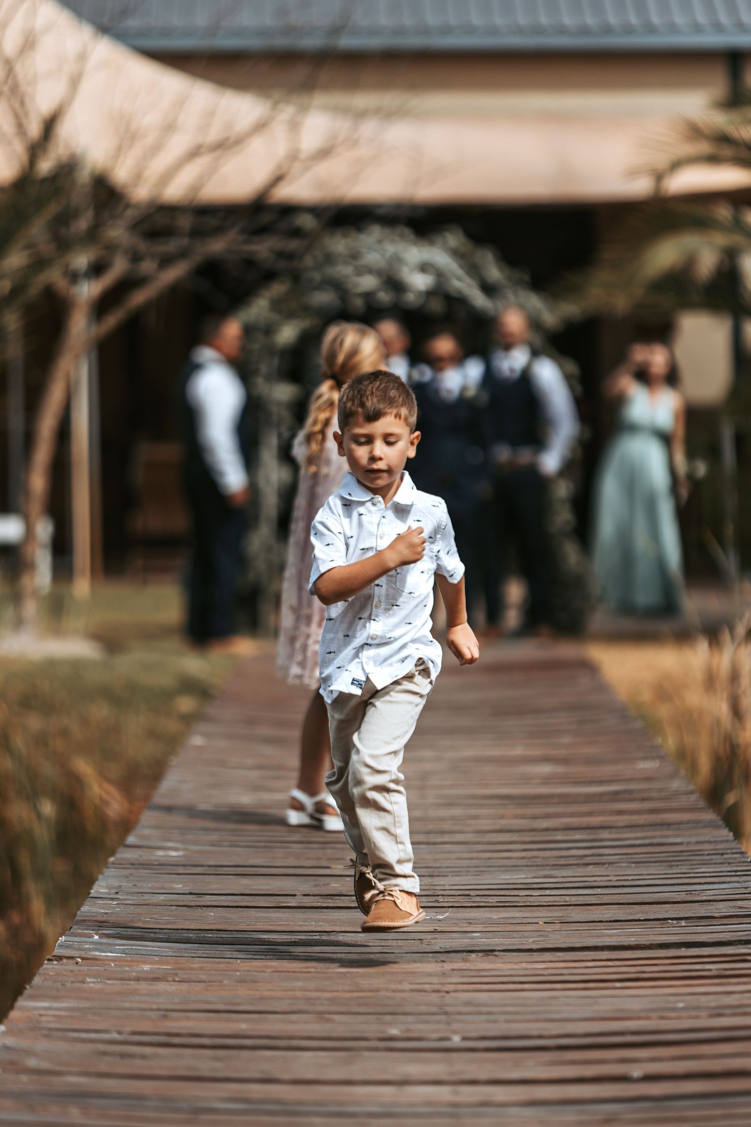 Young ring bearer runs energetically across wooden boardwalk during wedding celebration.