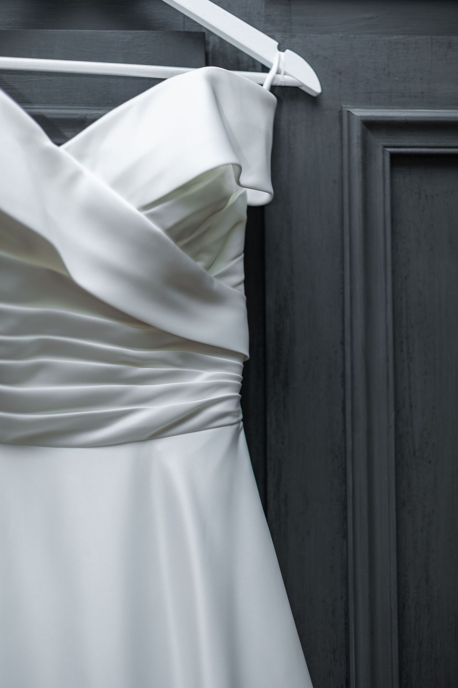 Elegant white off-shoulder wedding dress hanging on hanger against dark door.