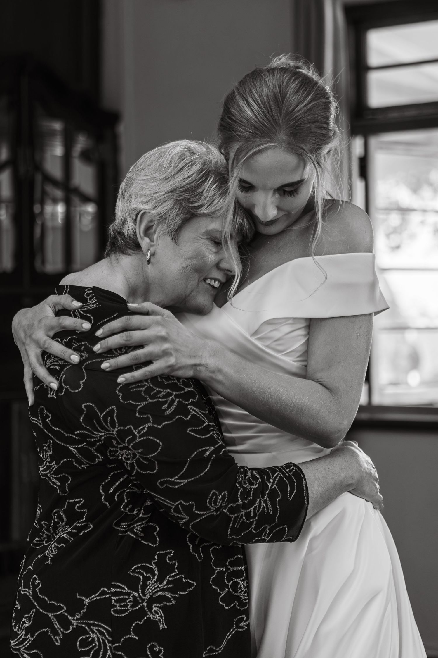 Emotional black and white photo of a bride in an off-shoulder gown sharing a tender hug with an older woman.