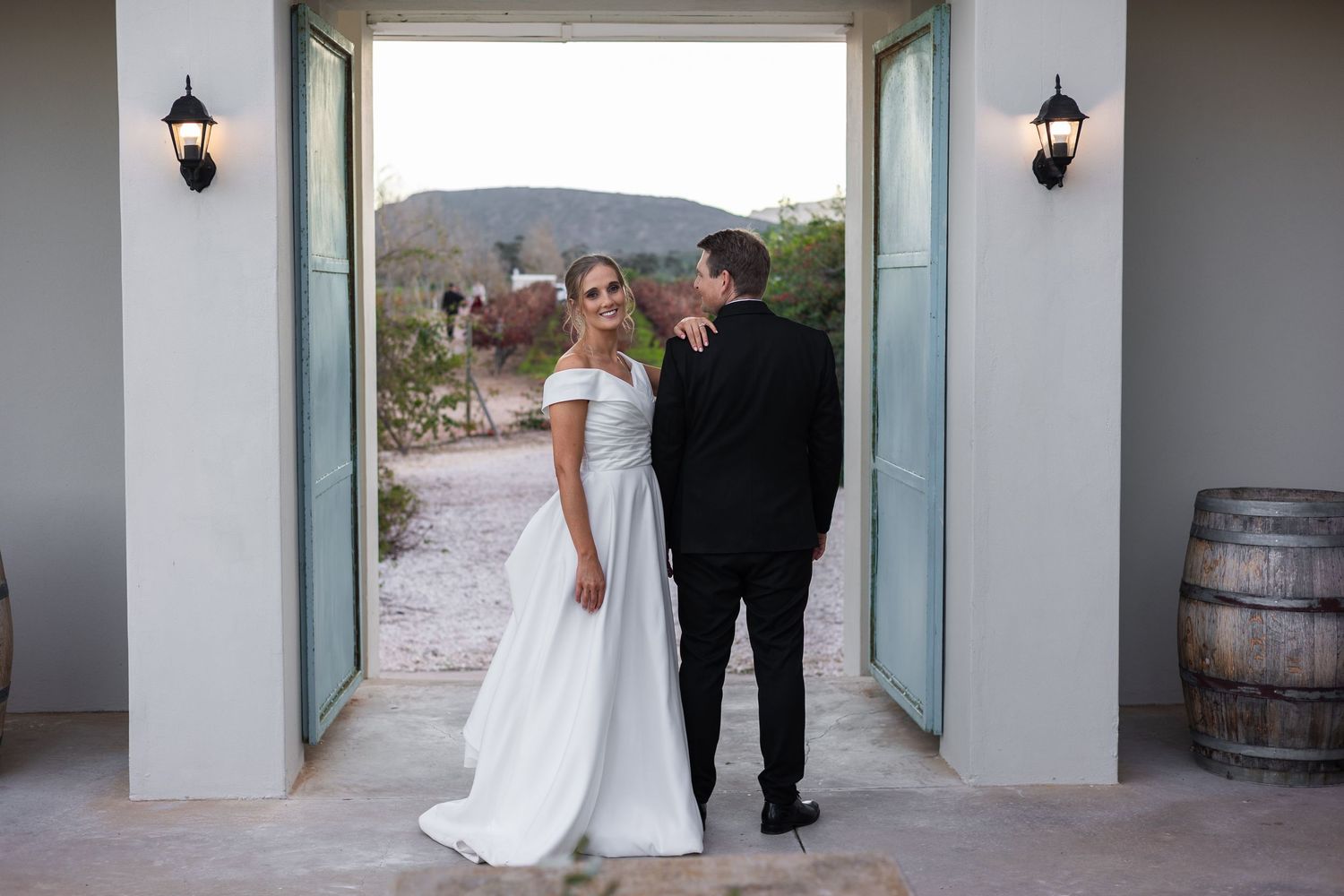 Bride in white off-shoulder gown and groom in black suit stand in open doorway with mountain views at outdoor wedding venue.