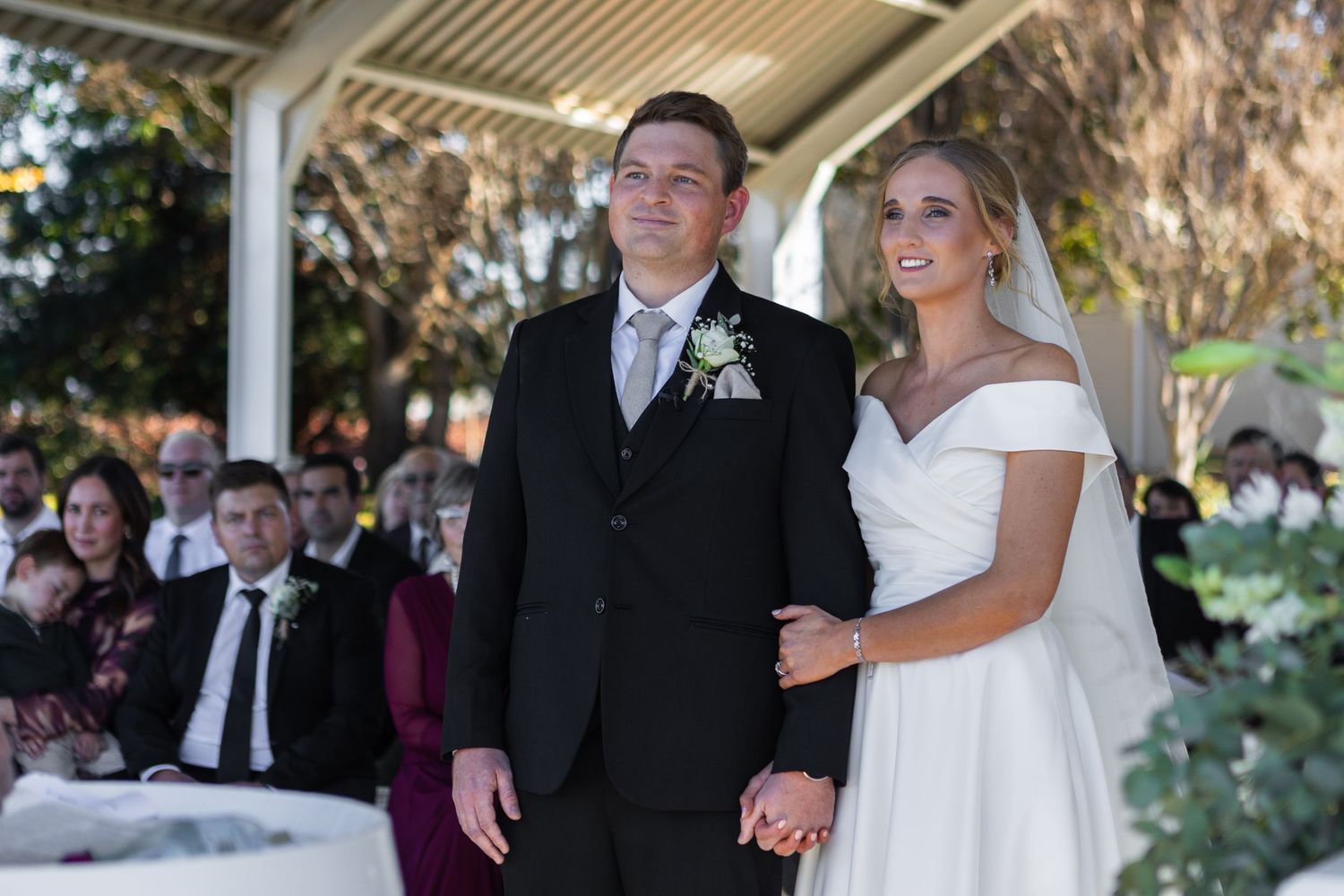 Bride in off-shoulder gown and groom in black suit stand together at outdoor ceremony.