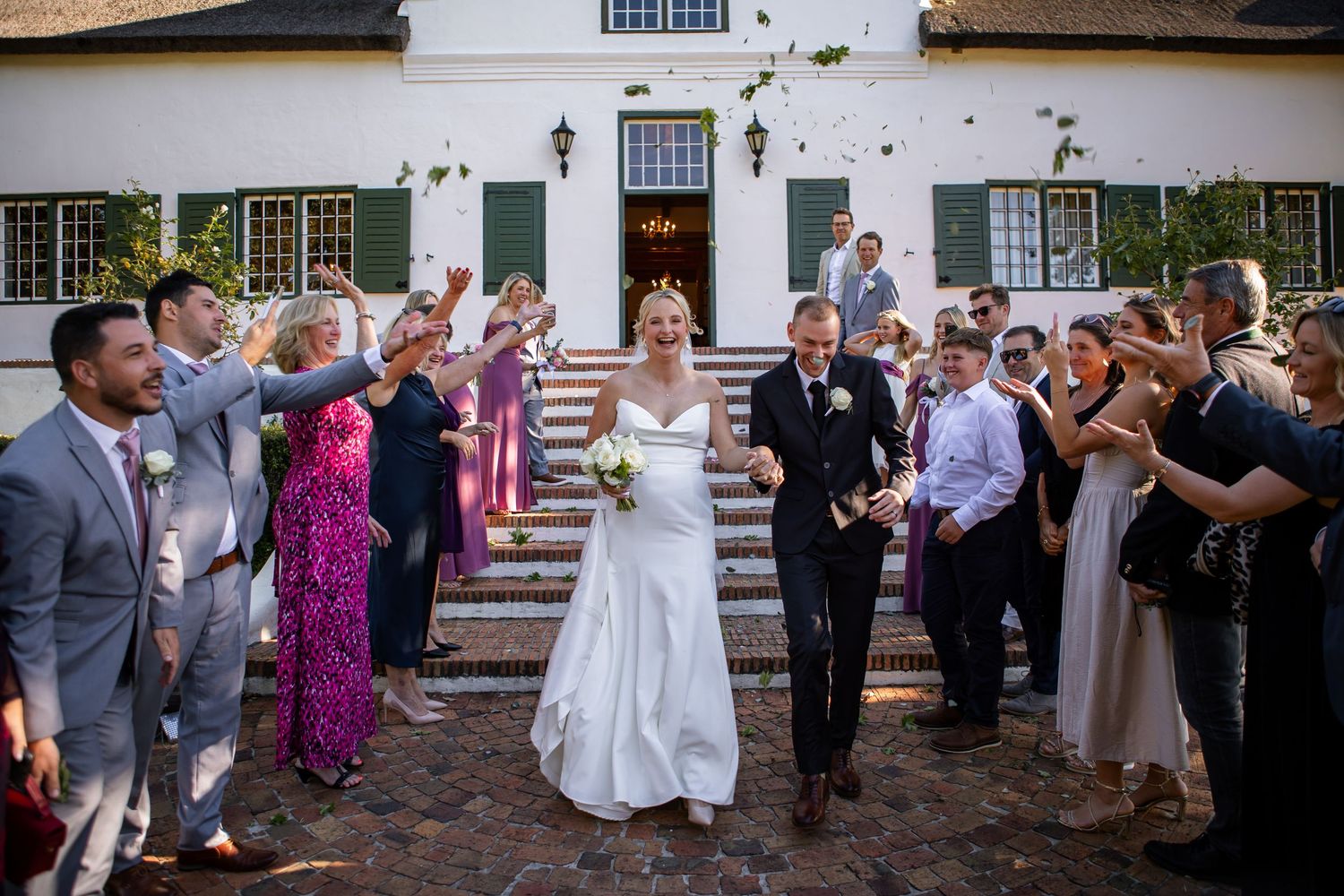 Wedding party and guests line up outside a white colonial building for a celebratory moment.