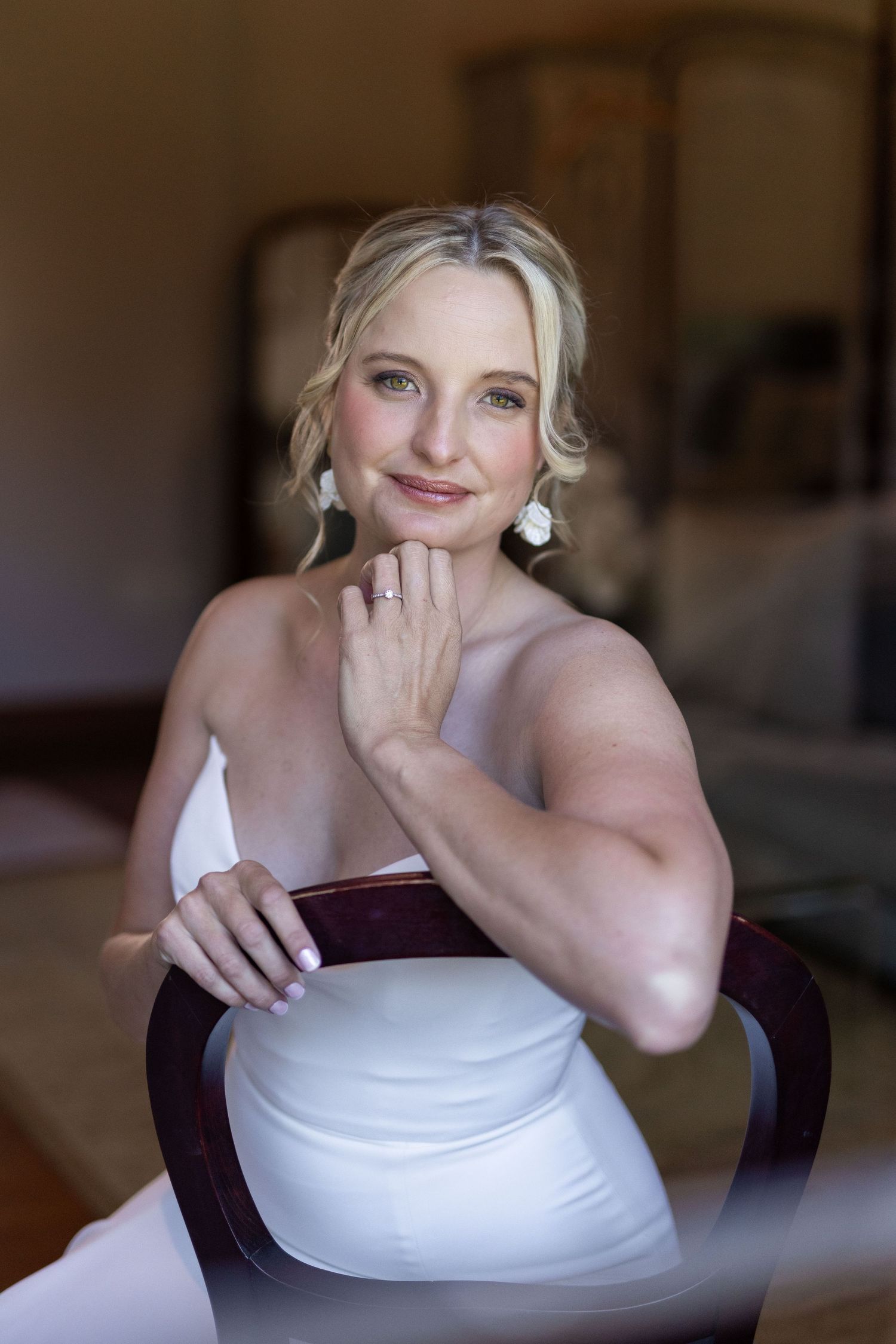 A woman in a white dress poses elegantly in a chair while displaying jewelry and a sophisticated updo hairstyle.