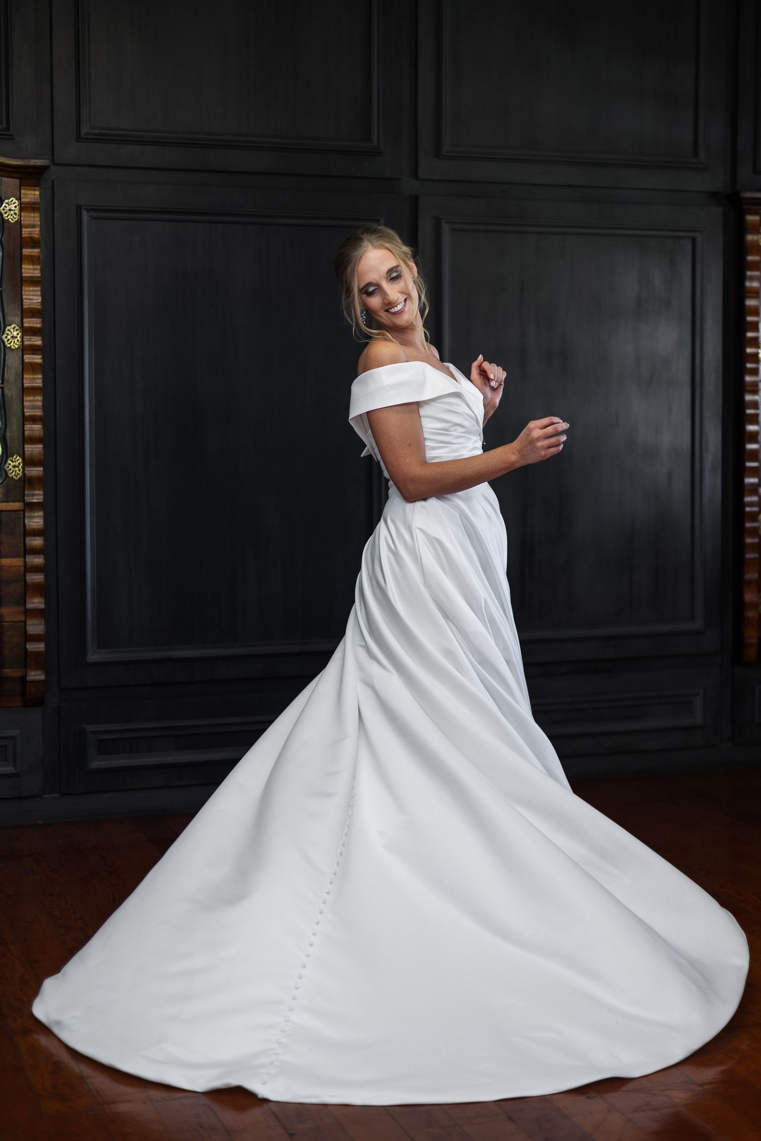 Bride in elegant off-shoulder white satin ball gown with long train, smiling against dark wood paneled wall.