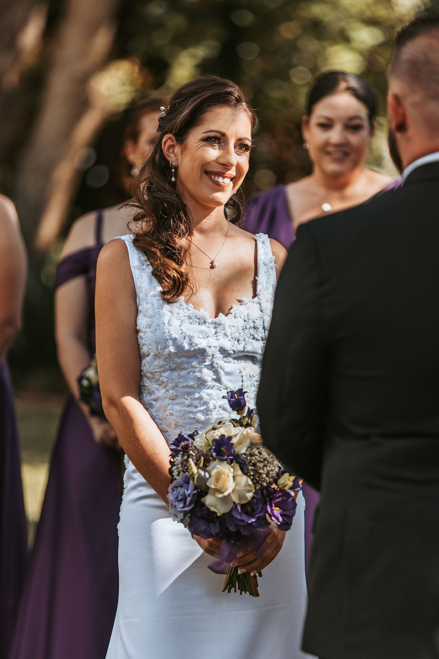 Bride in white lace dress smiles while holding purple and white bouquet during outdoor wedding ceremony.