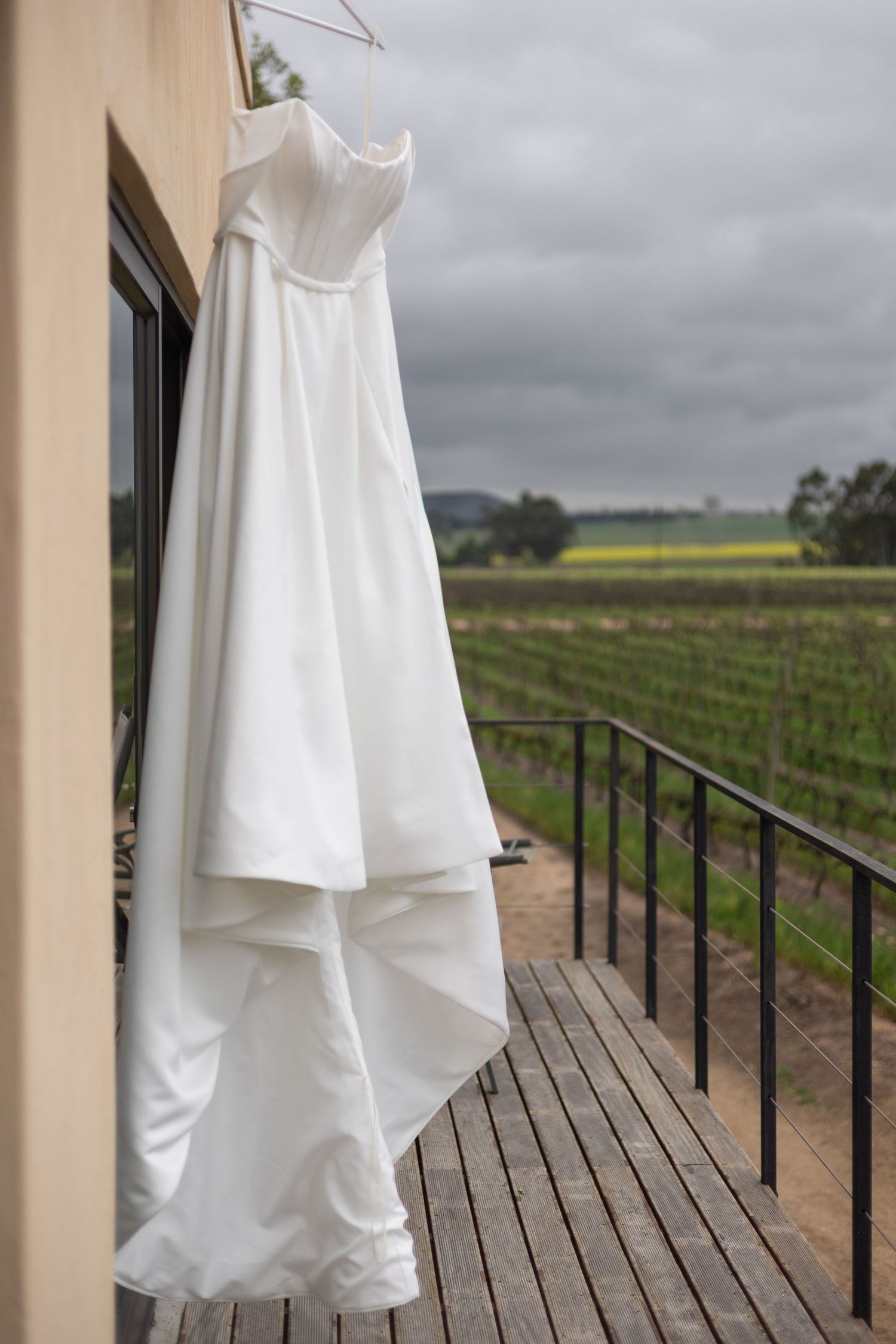 White strapless wedding dress hanging on a balcony railing with vineyard and cloudy sky in the background.