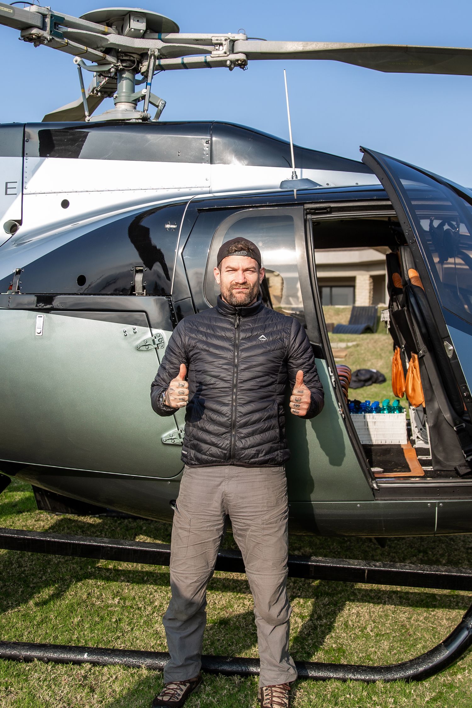 Bearded man in black puffer jacket giving thumbs up in front of a dark green helicopter on a sunny day.