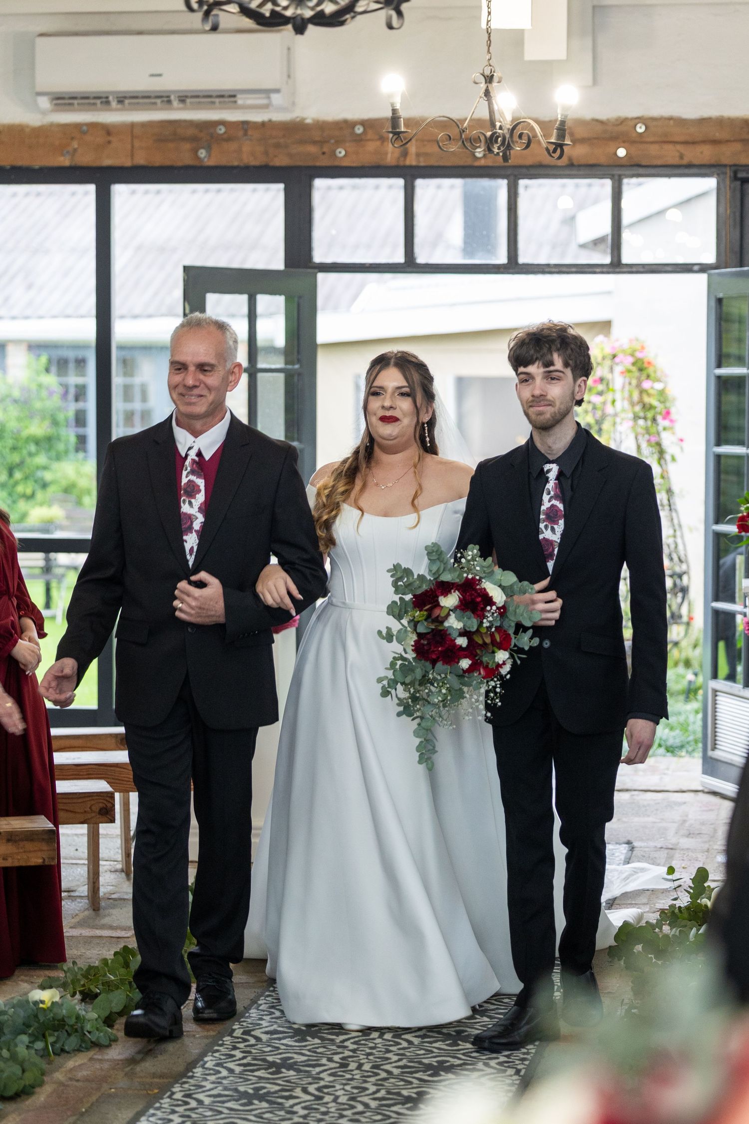 Bride walks down aisle escorted by two men in black suits with floral ties, holding red bouquet in elegant venue.