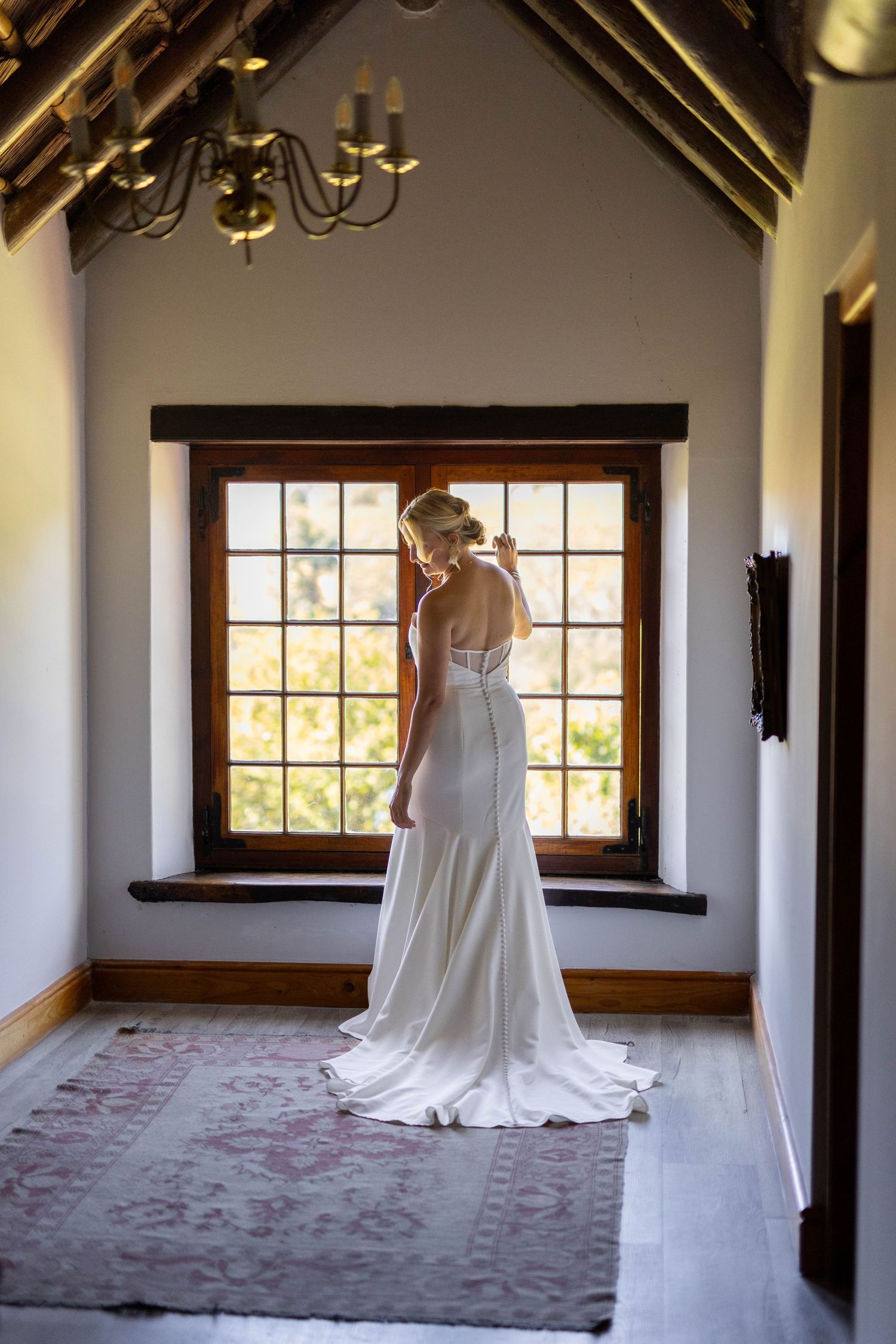 Elegant wedding dress with long train photographed in sunlit room with wooden beams and windows.