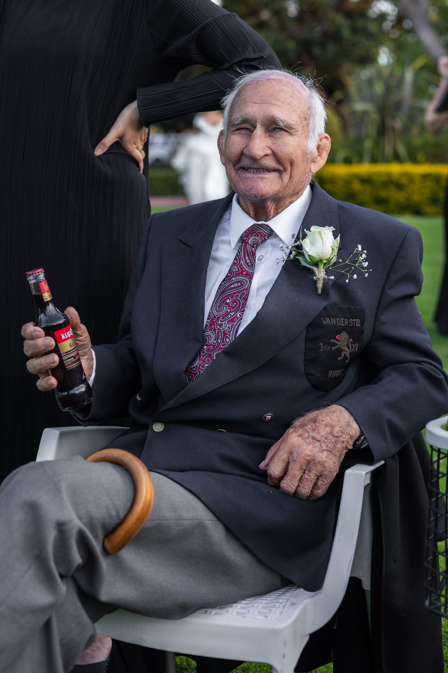 Elderly man in suit with boutonniere smiles while holding a bottle at an outdoor wedding.