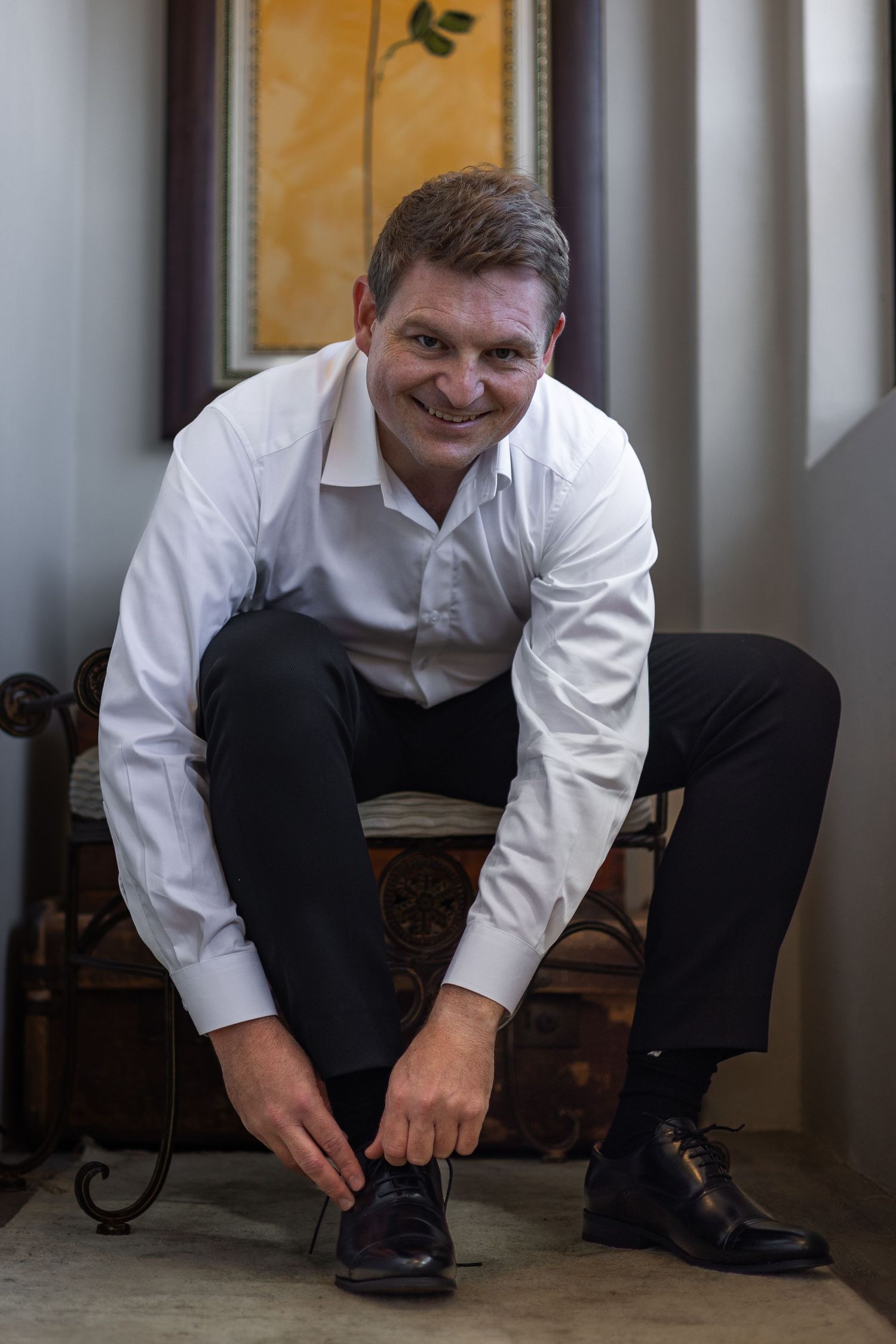 Smiling groom in white shirt tying his black dress shoes while seated on a decorative chair before his wedding.