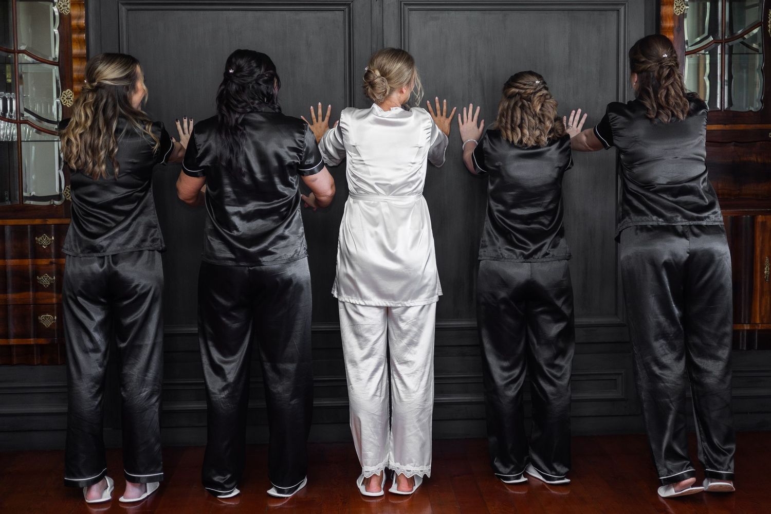 Bride in white satin pajamas with four bridesmaids in black satin pajamas, all facing a dark wall with hands pressed against it.