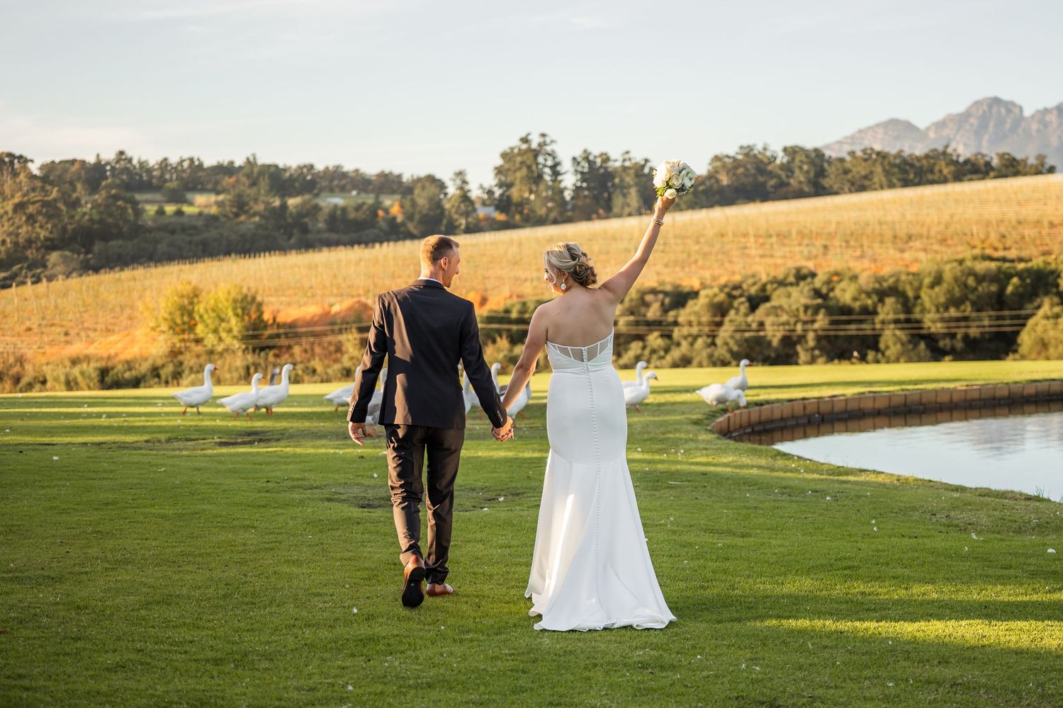 Newlyweds walk hand in hand across a scenic golf course with mountains and lake in background.