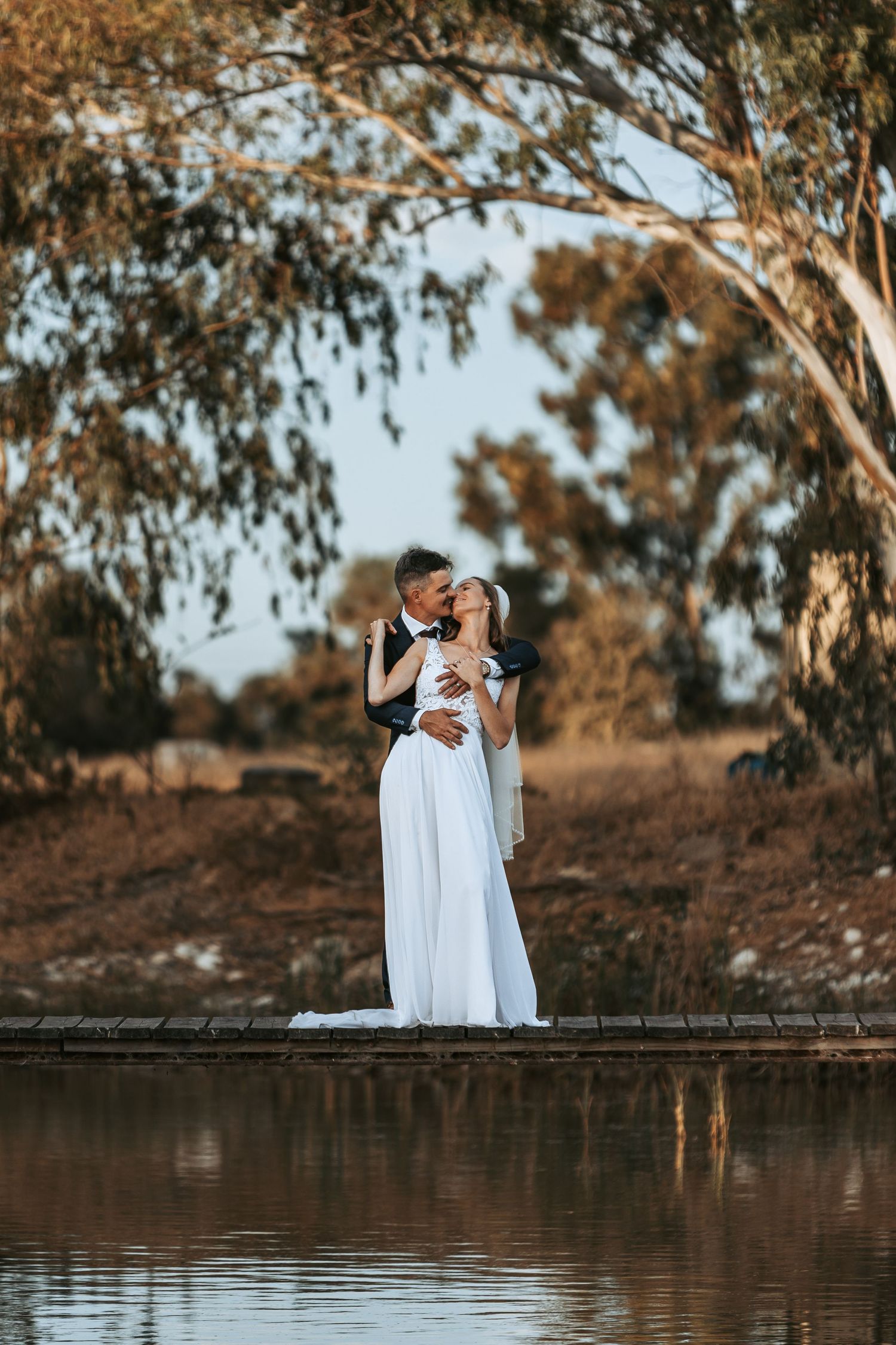 Wedding party poses on wooden boardwalk overlooking tranquil water at sunset.