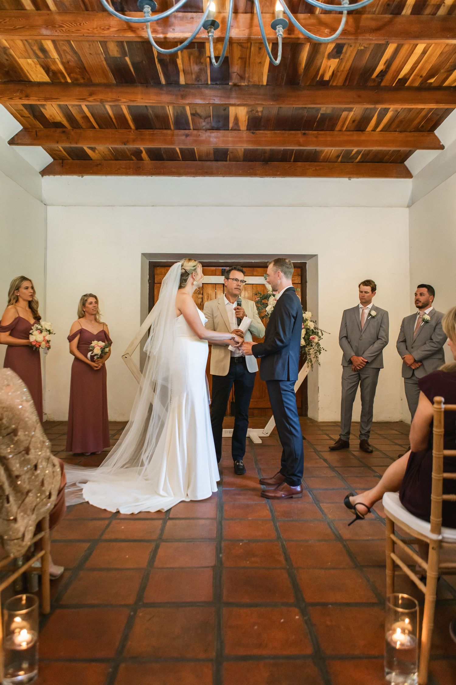 Indoor wedding ceremony with guests in formal attire on terracotta tile floor with wooden ceiling beams.