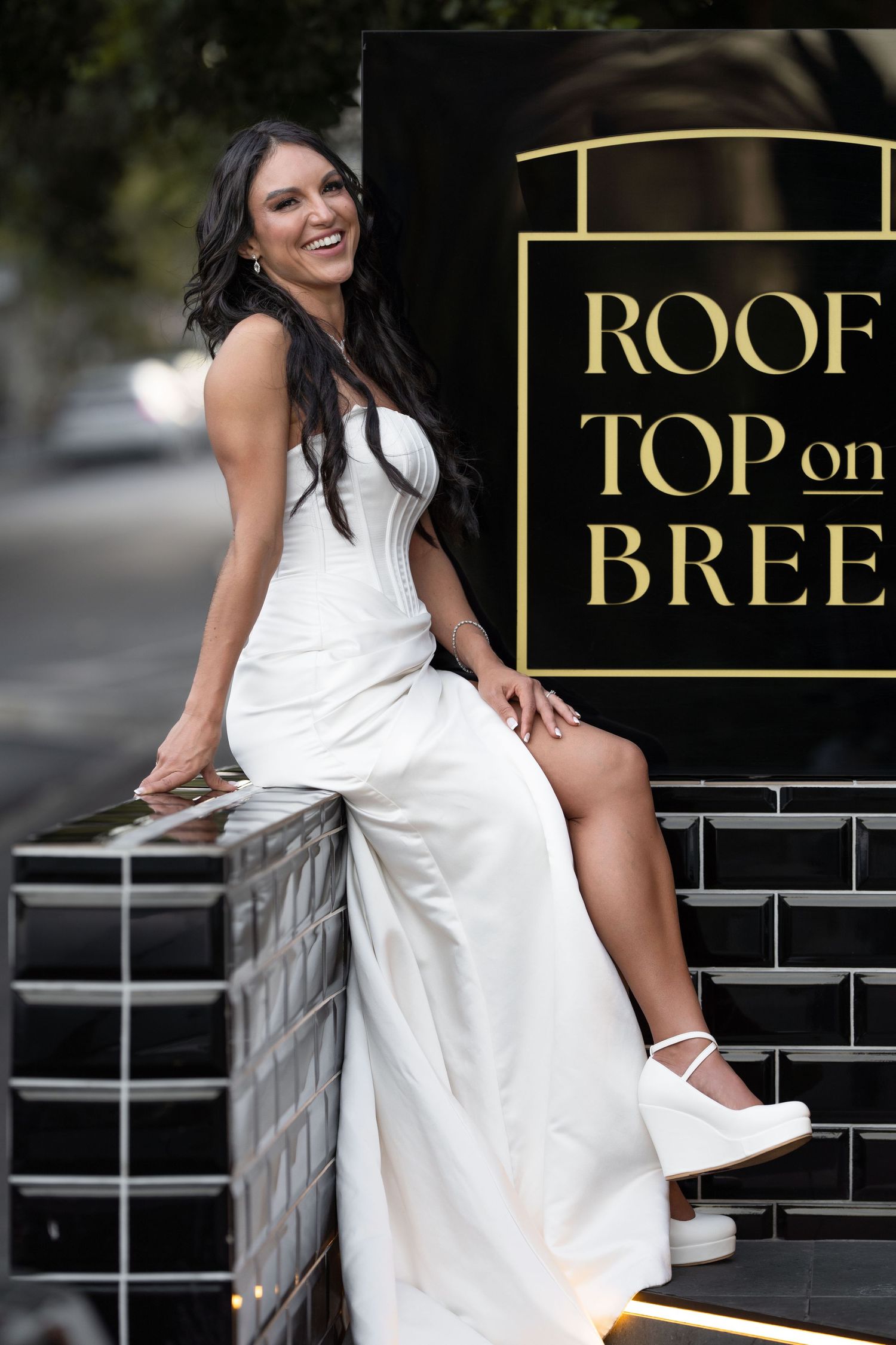 Woman in white dress posing at Rooftop on Bree venue entrance, smiling confidently.