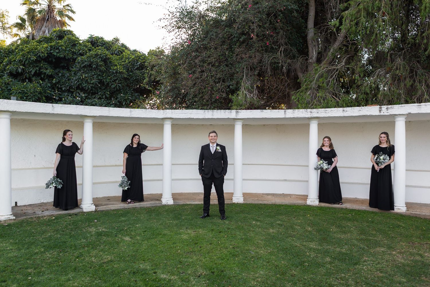 Groom in black suit stands with four bridesmaids in black dresses at outdoor wedding venue.
