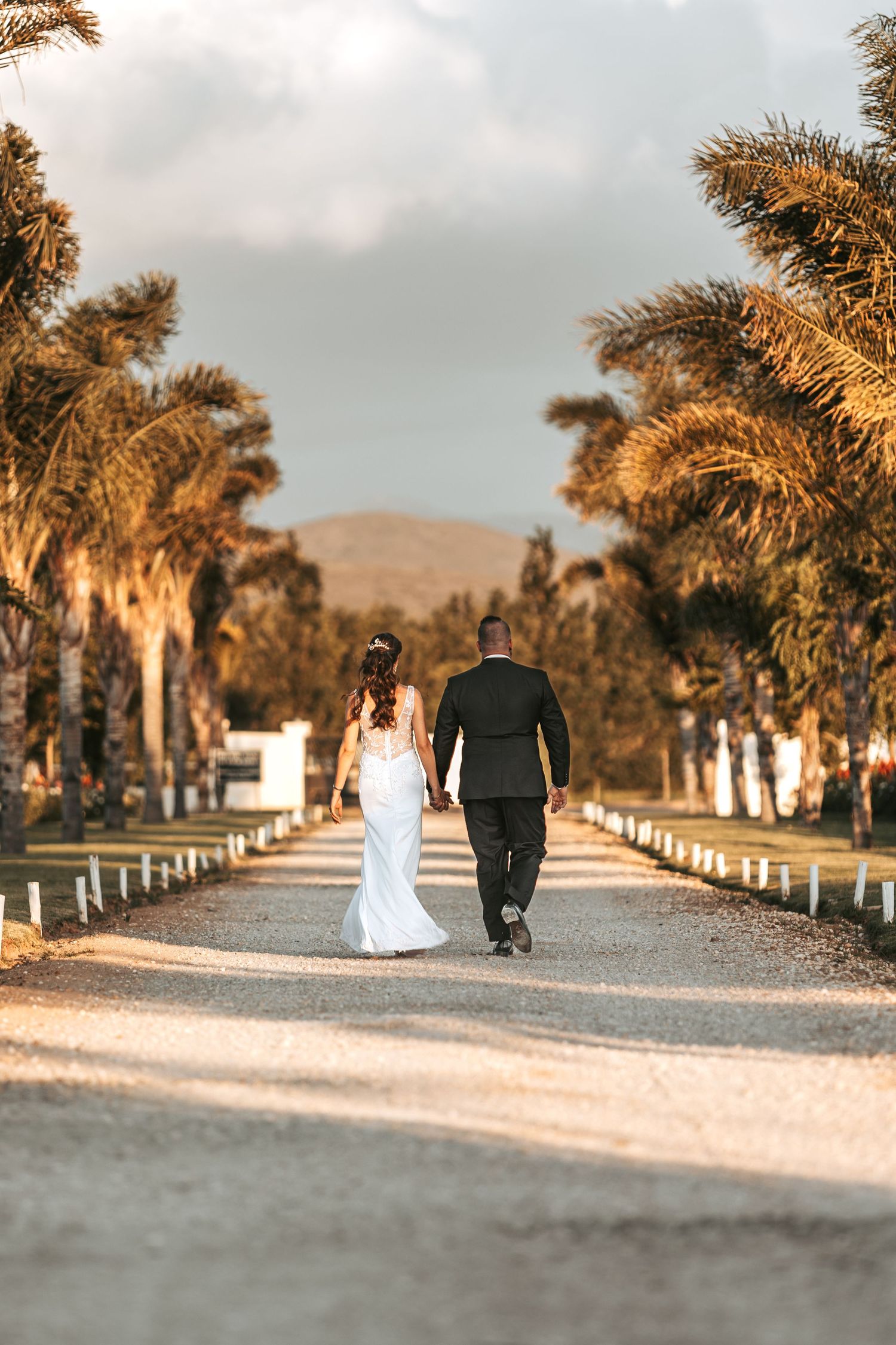 Newlyweds walk down palm tree-lined path at sunset after wedding ceremony.