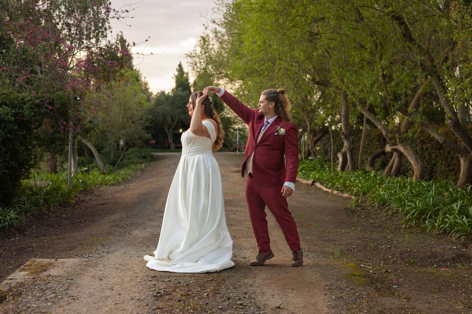 Bride and groom dancing on a tree-lined path, groom in burgundy suit twirling bride in white gown outdoors.
