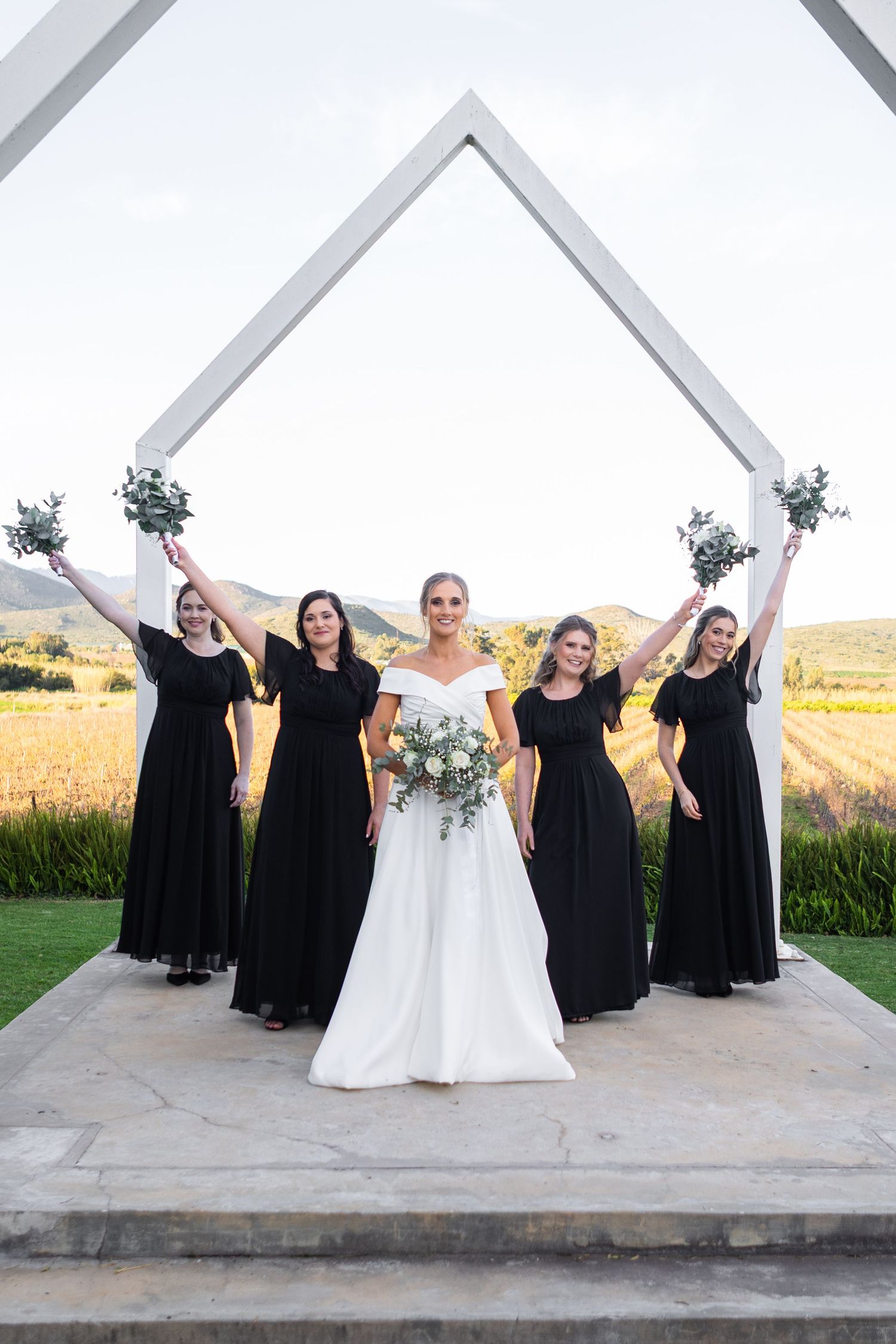 Bride in white gown with four bridesmaids in black dresses celebrating under white arch.