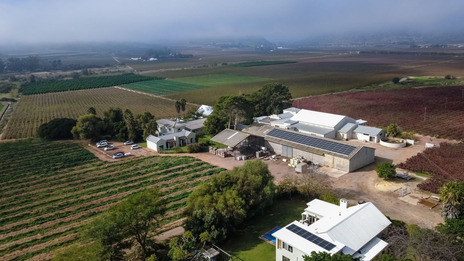 Aerial view of a winery estate with vineyards, farm buildings, and rolling green fields under a cloudy sky.