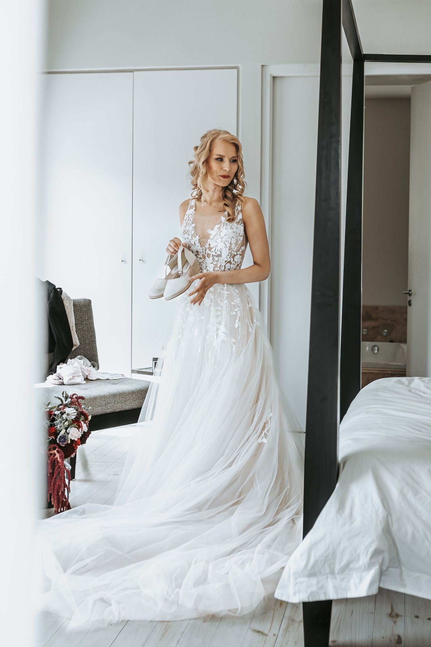 Full-length shot of bride in white lace wedding dress with flowing train standing in bedroom.