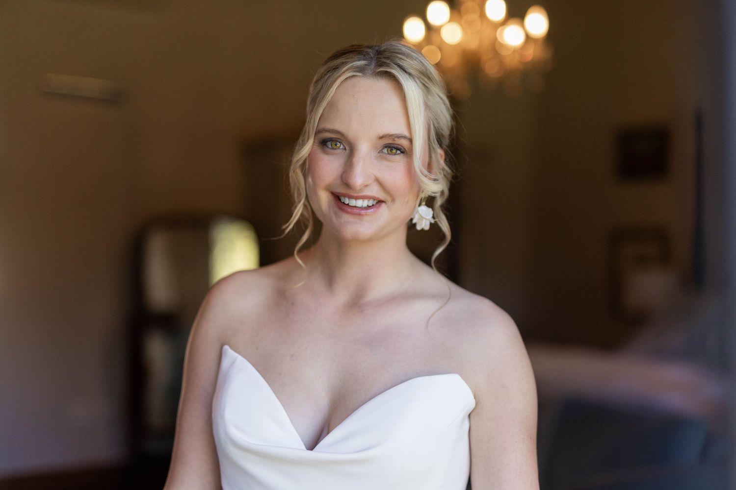 A bride in a white strapless wedding gown smiles while posing for photos in an elegant indoor setting.