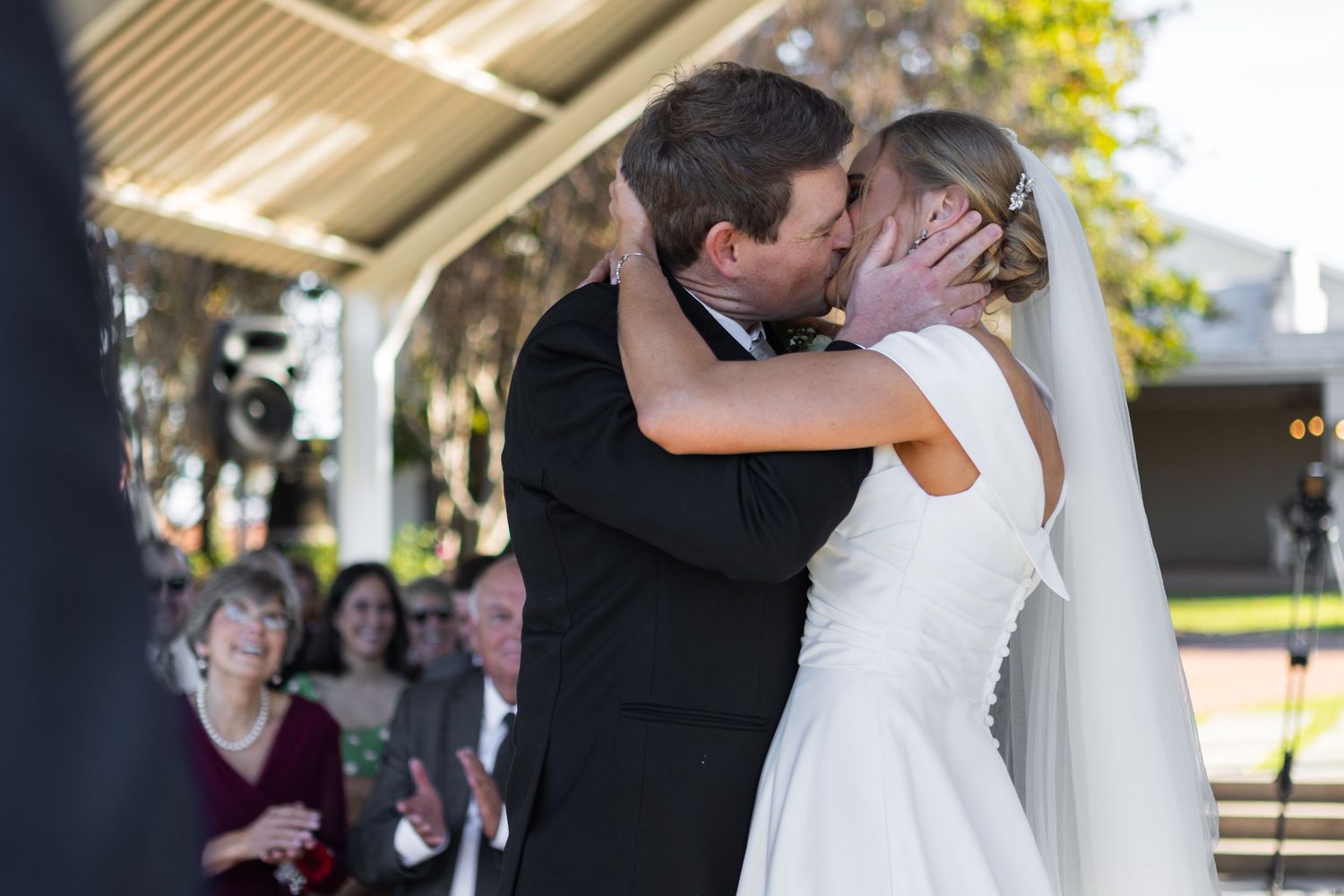 Bride and groom sharing their first kiss at the altar surrounded by smiling wedding guests at the ceremony.