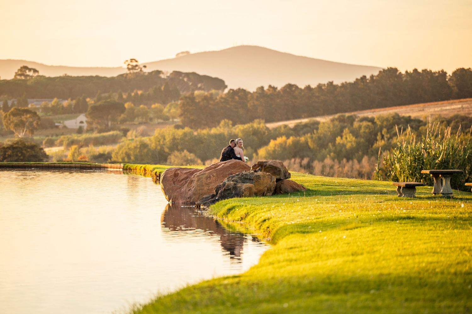A peaceful lakeside scene at sunset with someone sitting on a rock overlooking the water and rolling hills in the background.