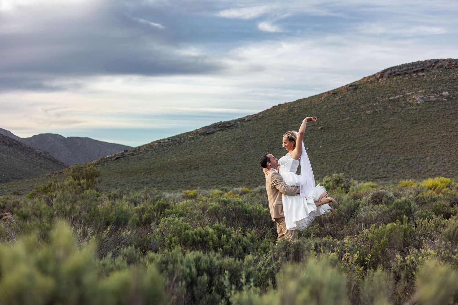 Couple dancing in lush green mountain landscape, man lifting woman in white dress under cloudy sky.