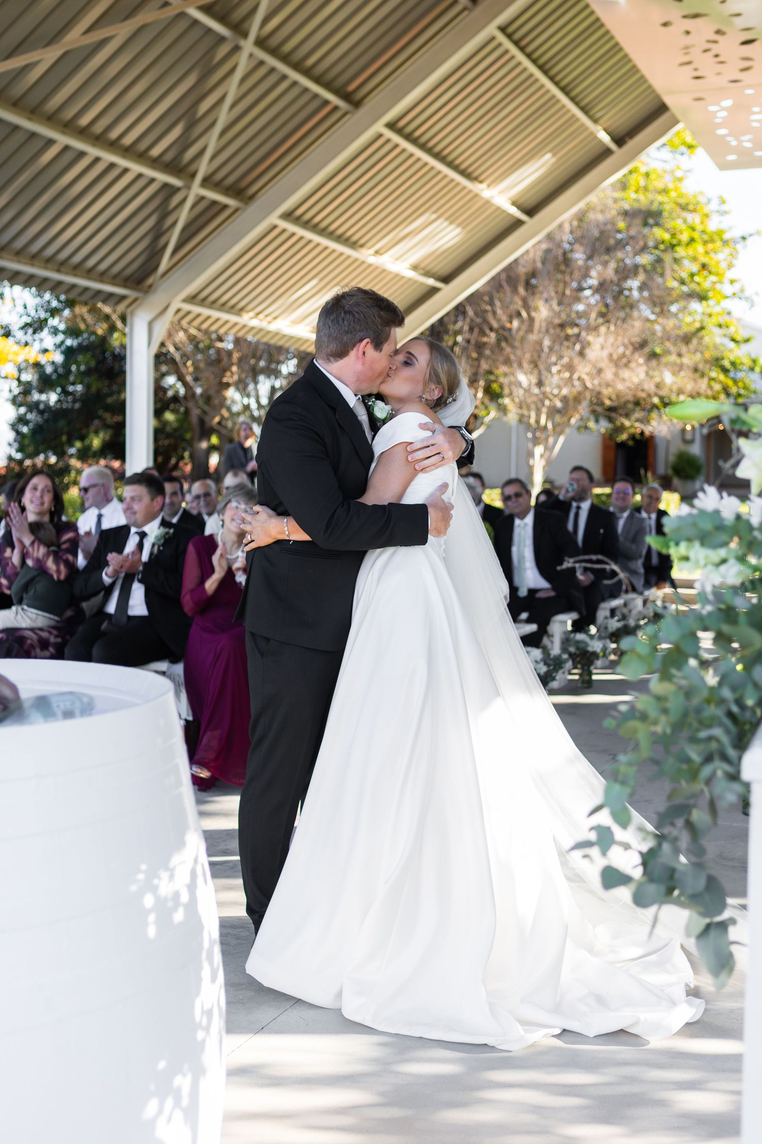 Bride and groom share first kiss under pavilion while wedding guests applaud in background.